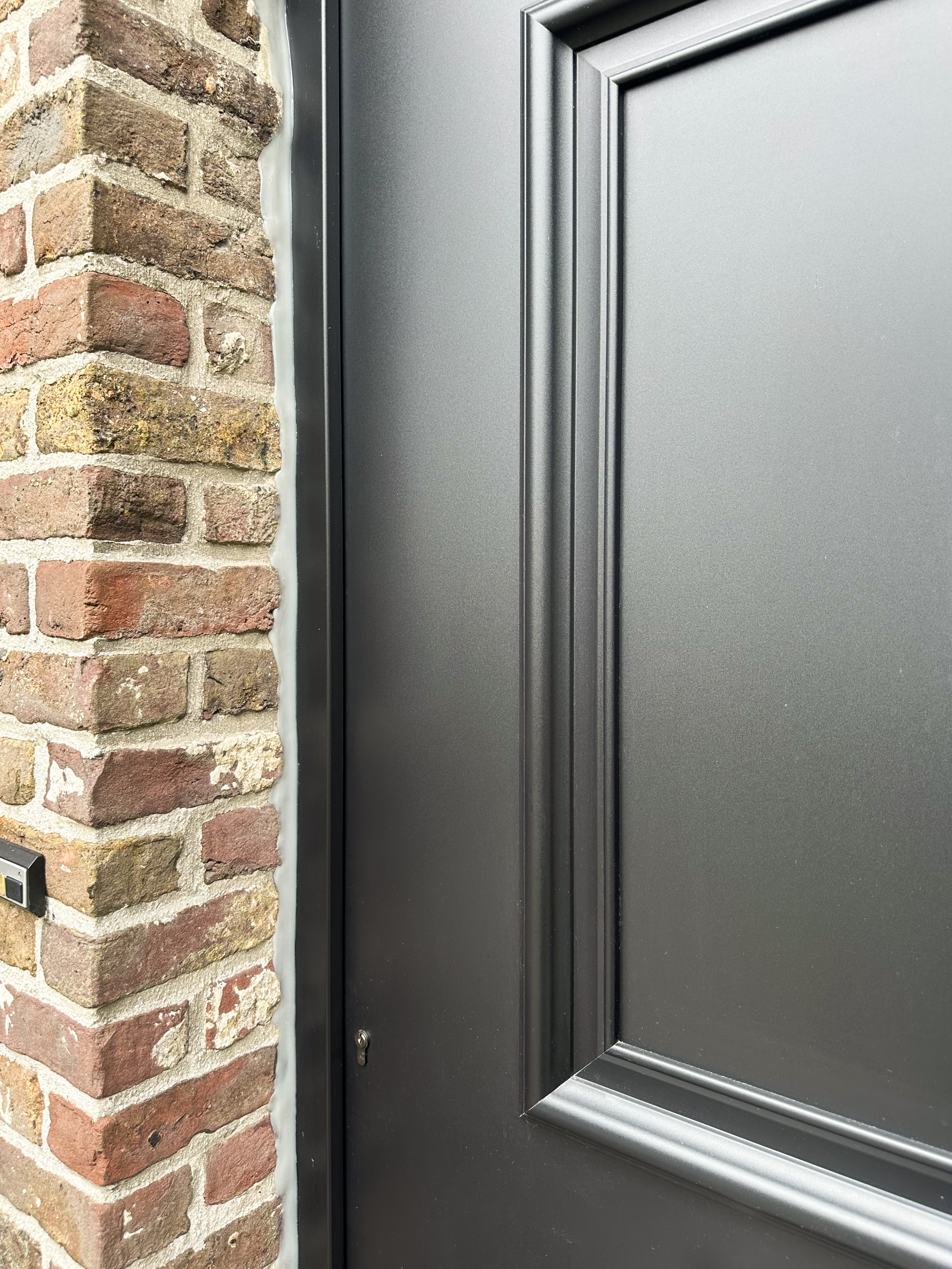 Detail van een matzwart PVC Impakt PLUS-paneel met decoratieve randprofilering, naast een gevel in zichtbare baksteen.
