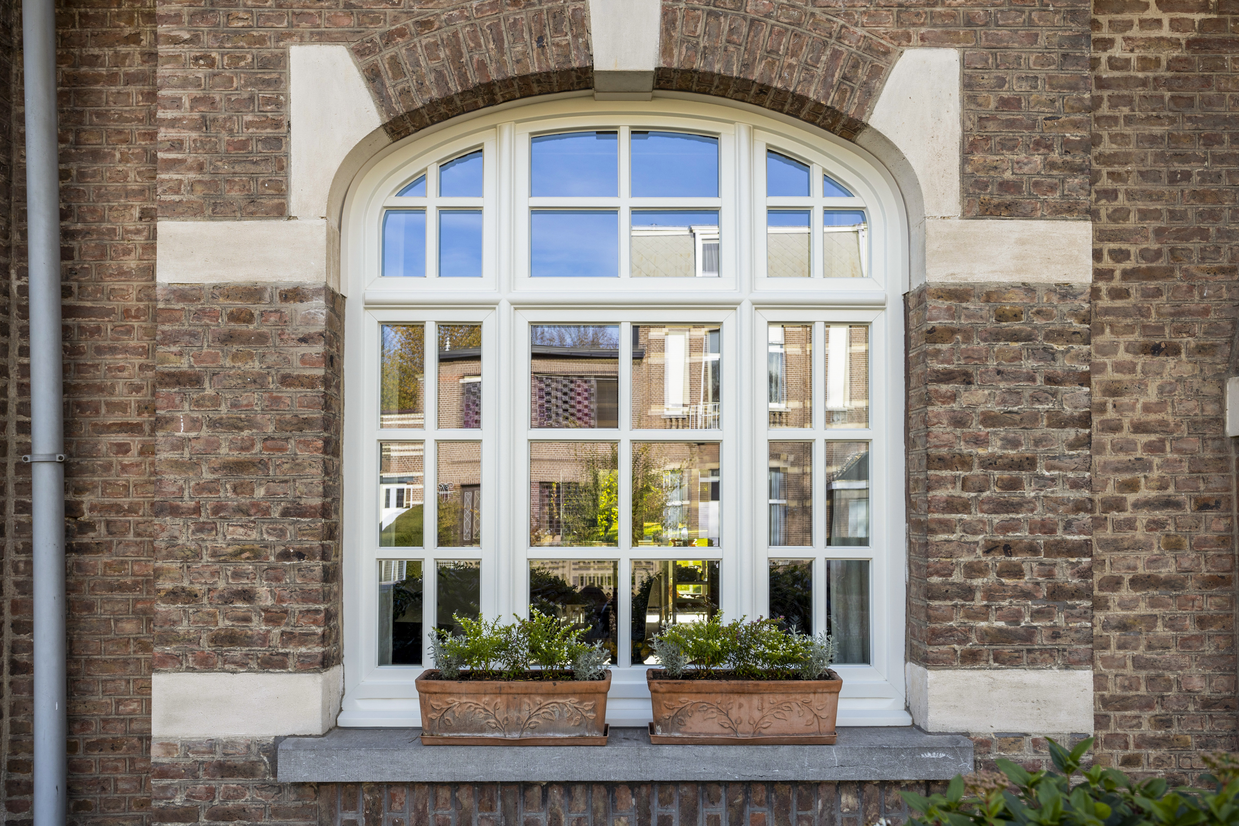 Wit afgelakt raam met drie verticale delen en bovenlicht in een historische bakstenen gevel met rondboog.