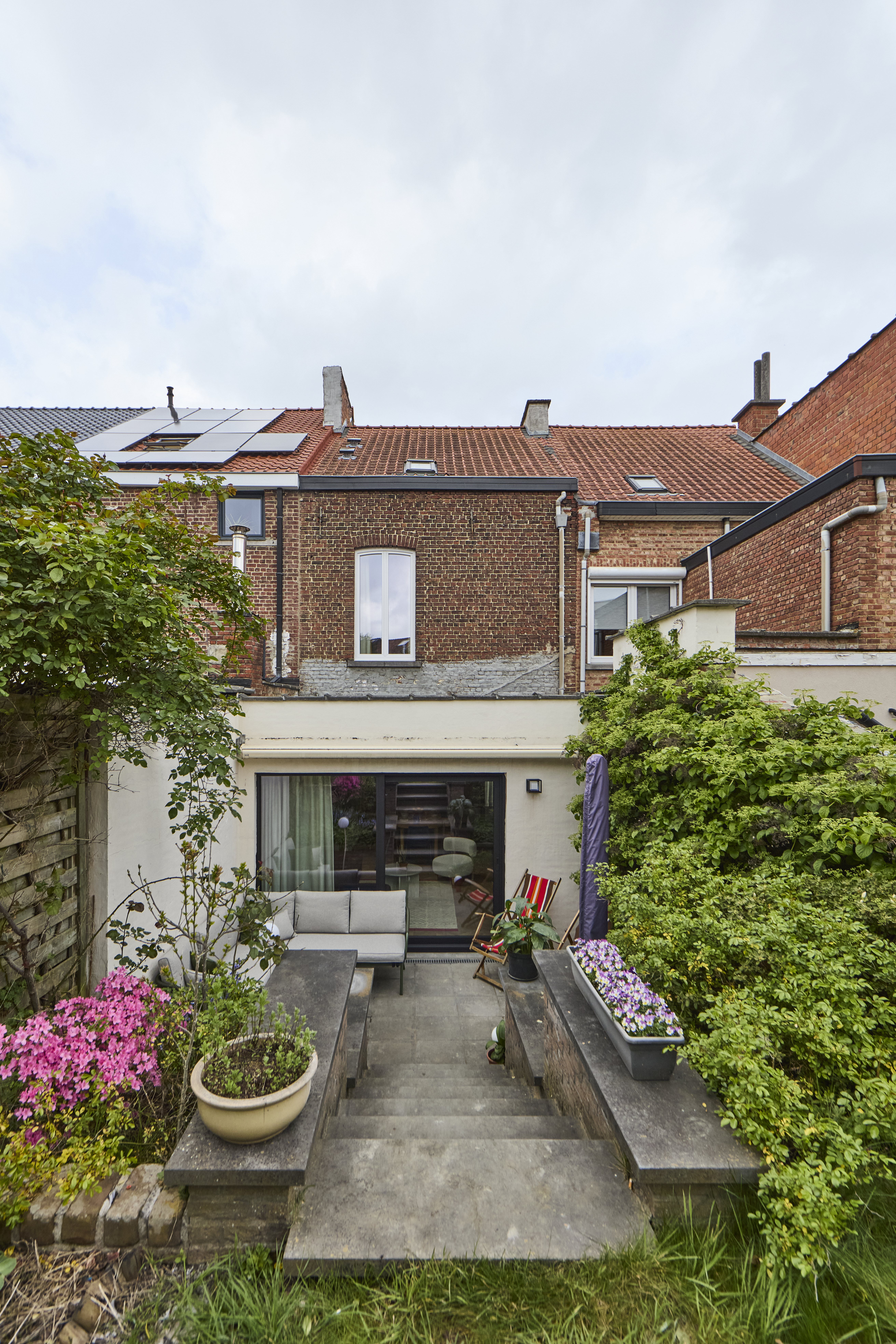 Achtergevel van een bakstenen woning met zwart aluminium schuifraam, terras en verzorgde tuin met bloemen en struiken.