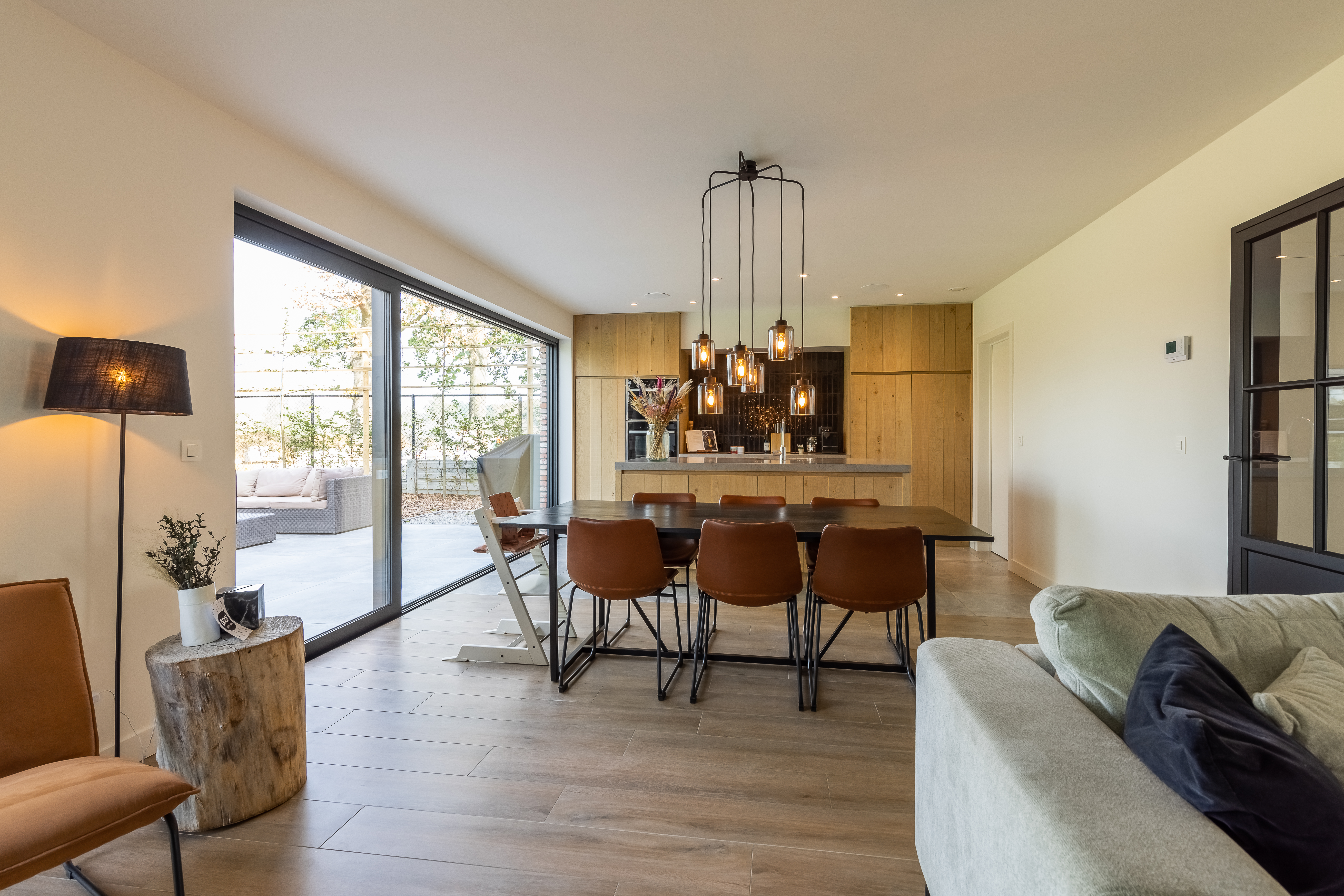 Warme, landelijke leefruimte in een nieuwbouwwoning met een diepzwart aluminium Visoglide Monorail-schuifraam, open keuken, houten vloer en zicht op het terras.