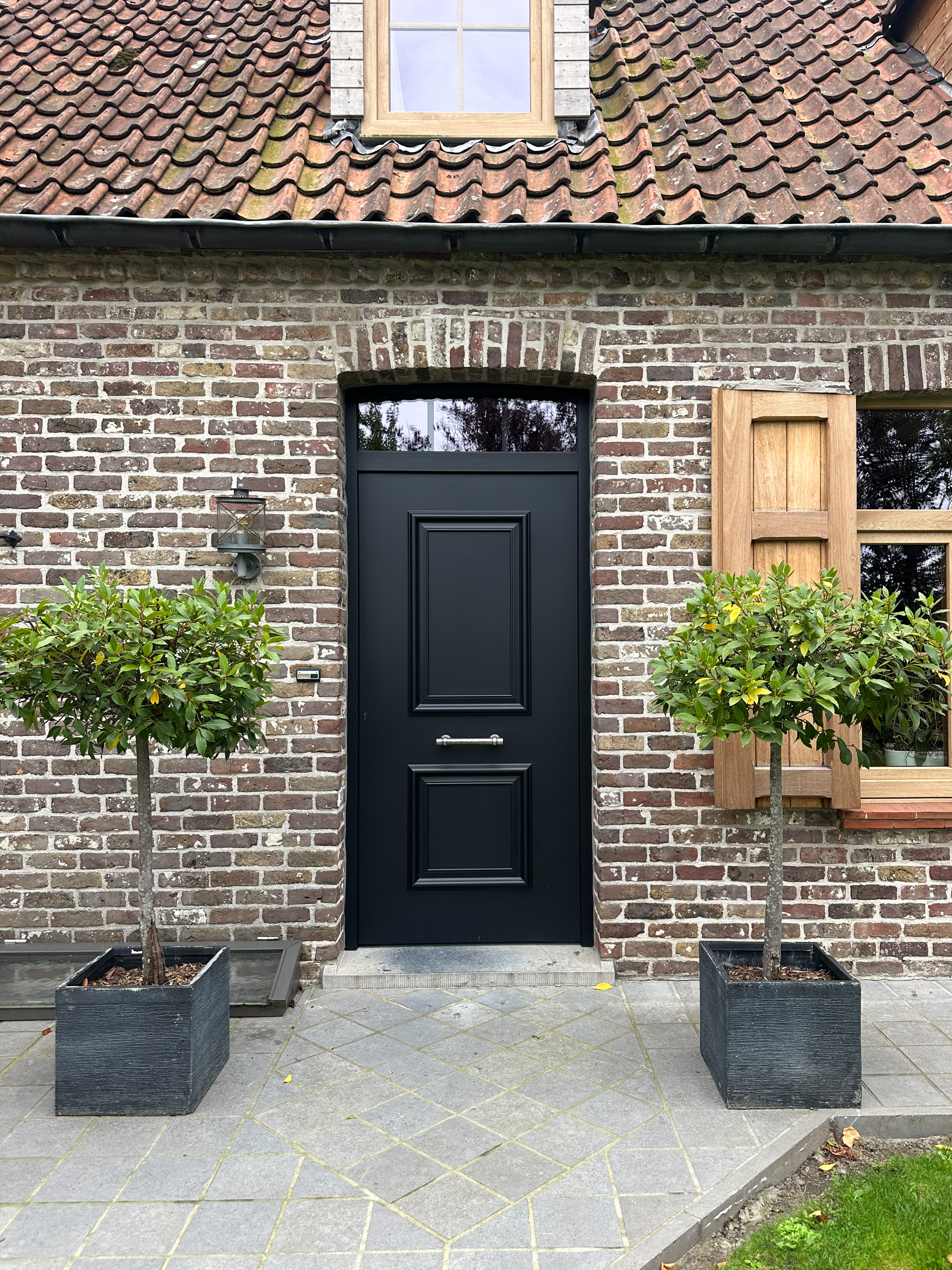 Matzwarte pvc-buitendeur met bovenlicht en deurpanelen in een bakstenen gevel, geflankeerd door twee plantenbakken.