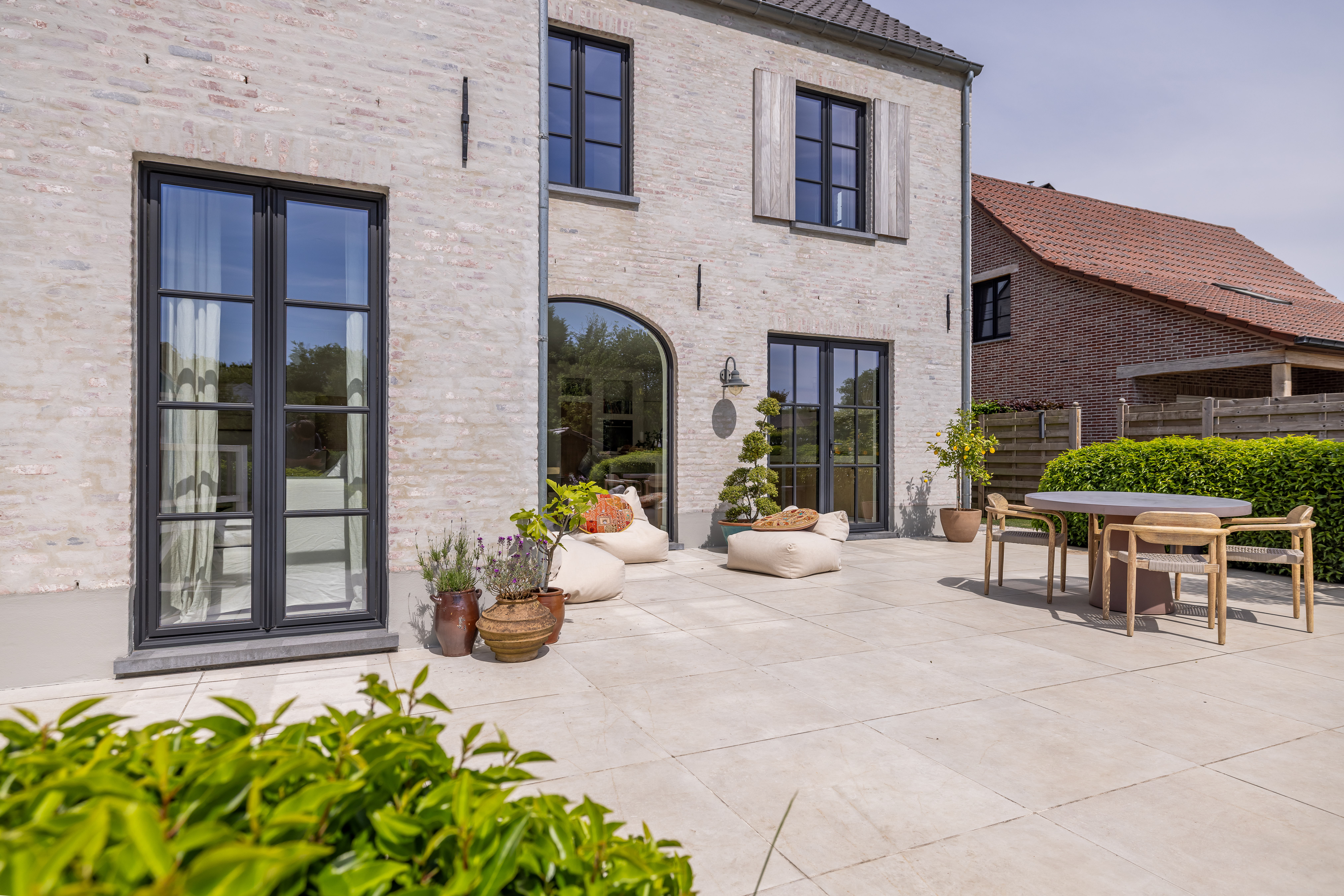 Lichte bakstenen gevel van een warm landelijke nieuwbouw met zwarte Steellook PLUS small-ramen, vaste ramen, dubbele ramen en draai-kipramen, aan een terras met tuinmeubelen.