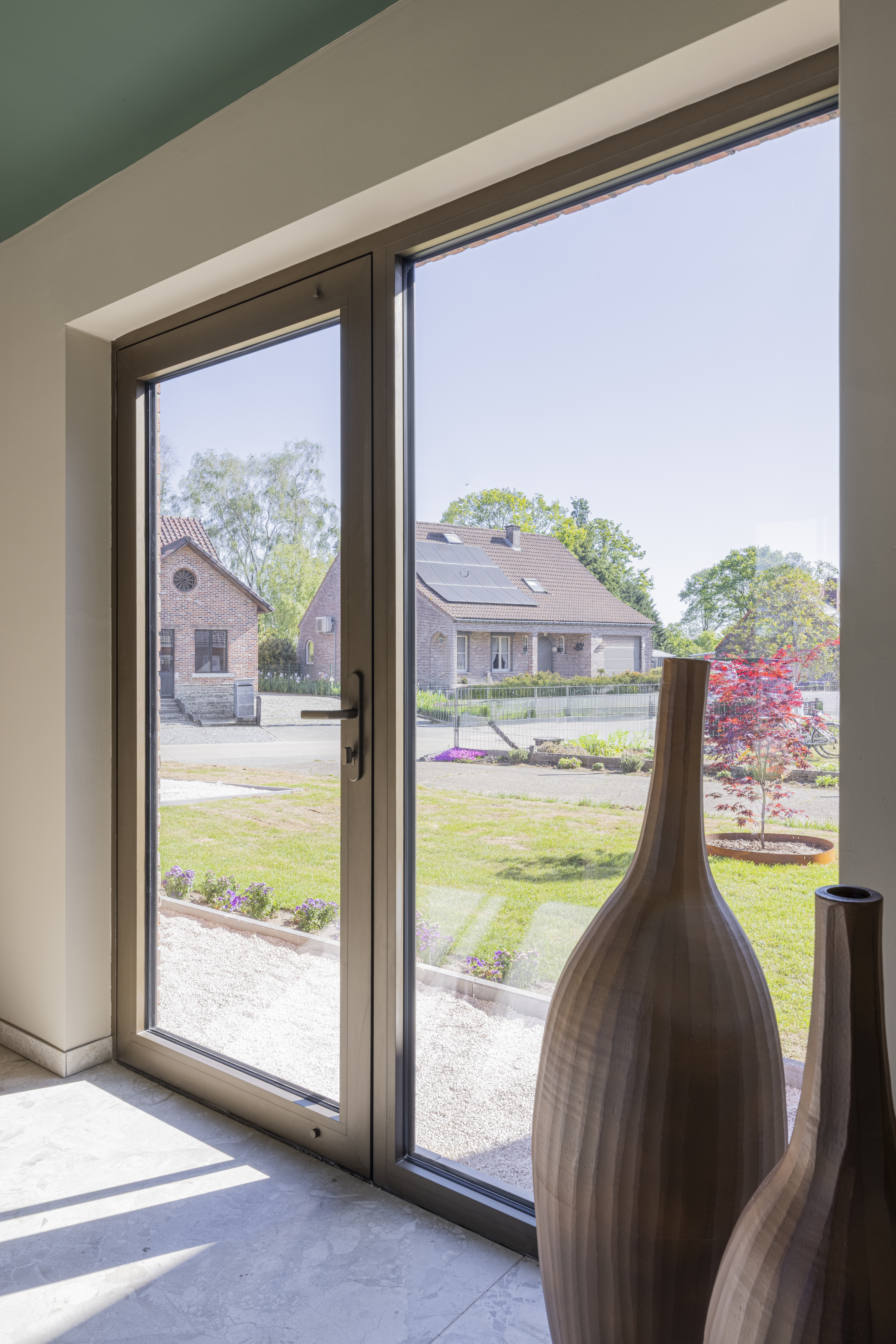 Beglaasde deur met een kamerhoog zijlicht in bronskleurig geanodiseerd aluminium, die vanuit een lichte binnenruimte uitkijkt op een groene voortuin.