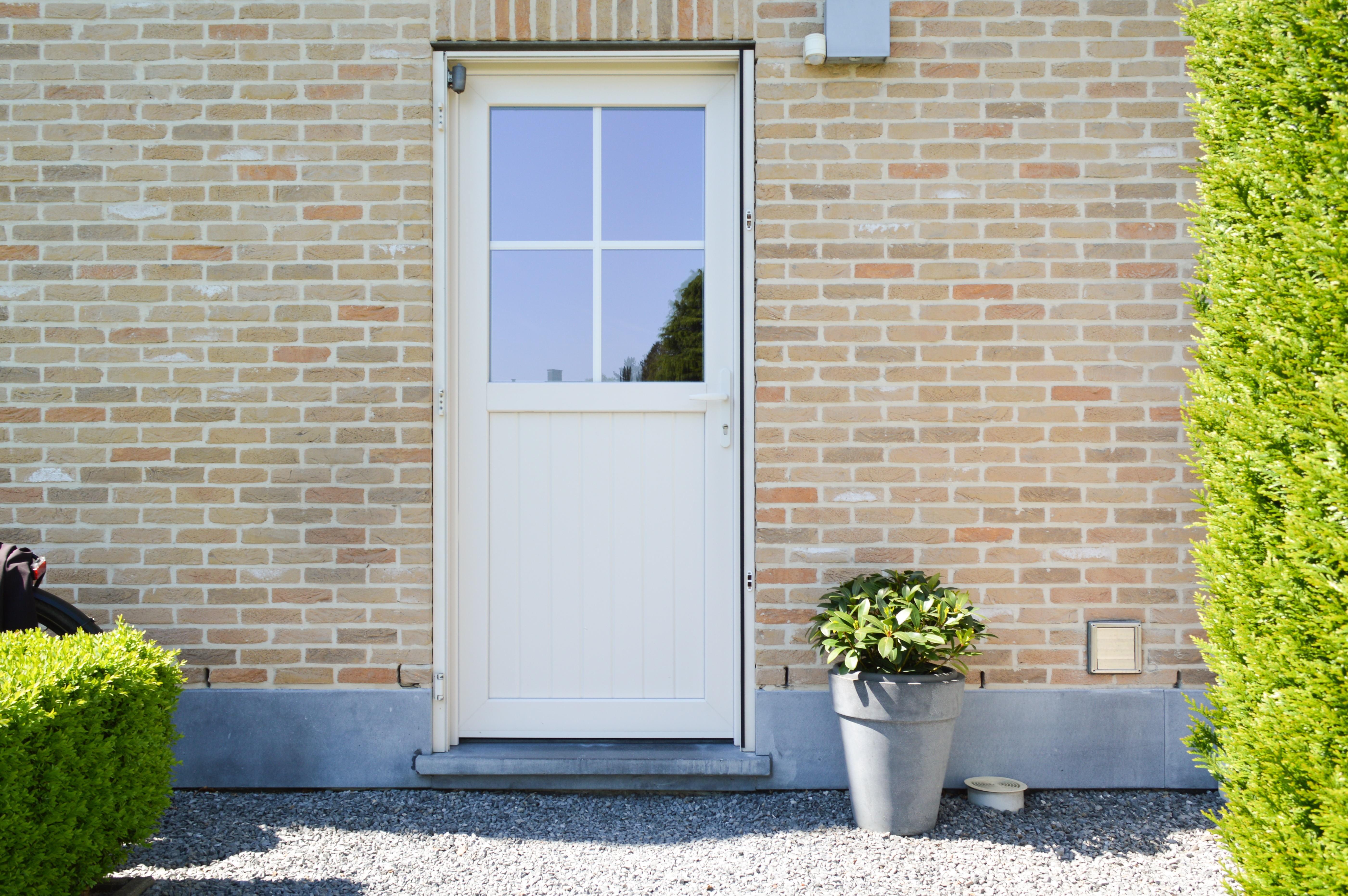Crèmekleurige, halfbeglaasde PVC-buitendeur uit de Impakt PLUS-reeks in een lichte bakstenen gevel, met wit raamprofiel, plantenpot en verzorgde voortuin.