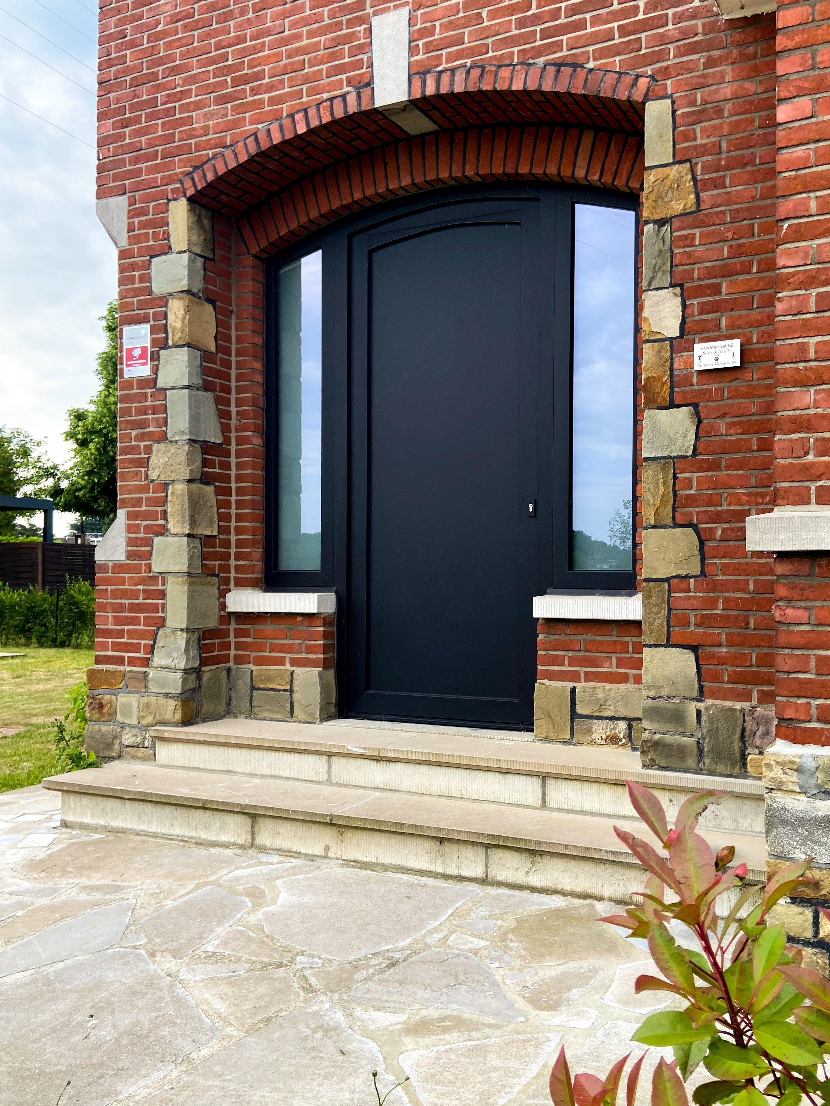 Gerenoveerde inkom van een vrijstaande woning met een matzwarte ALU Koncept-deur, smalle zijlichten en een bovenlicht, omlijst door rode bakstenen.