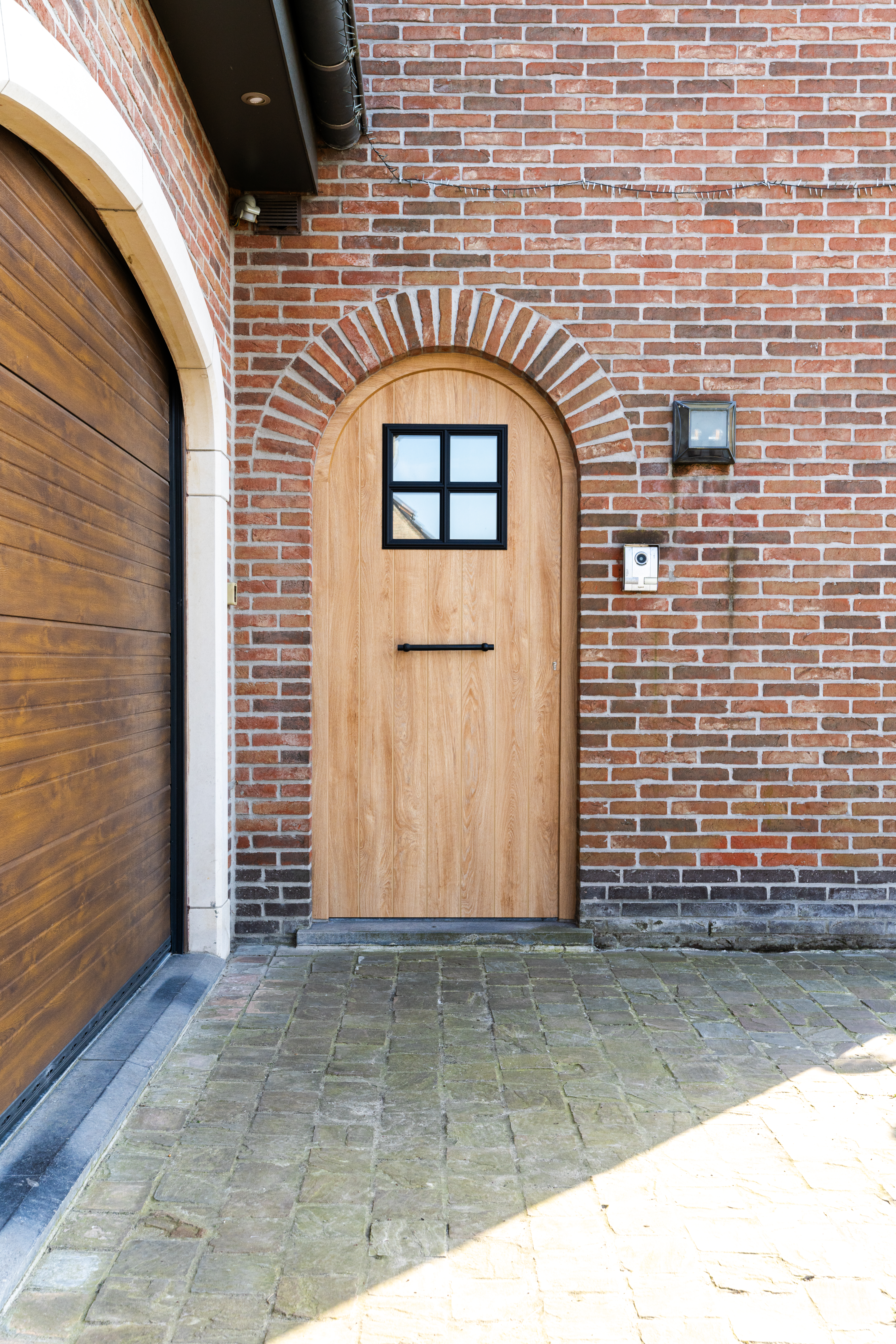 Bakstenen gevel met een voordeur in PVC IMPAKT PLUS met afwerking Woodec Eik Natuur en zichtbare nerf, naast een gebogen houten poort en een geplaveide oprit.