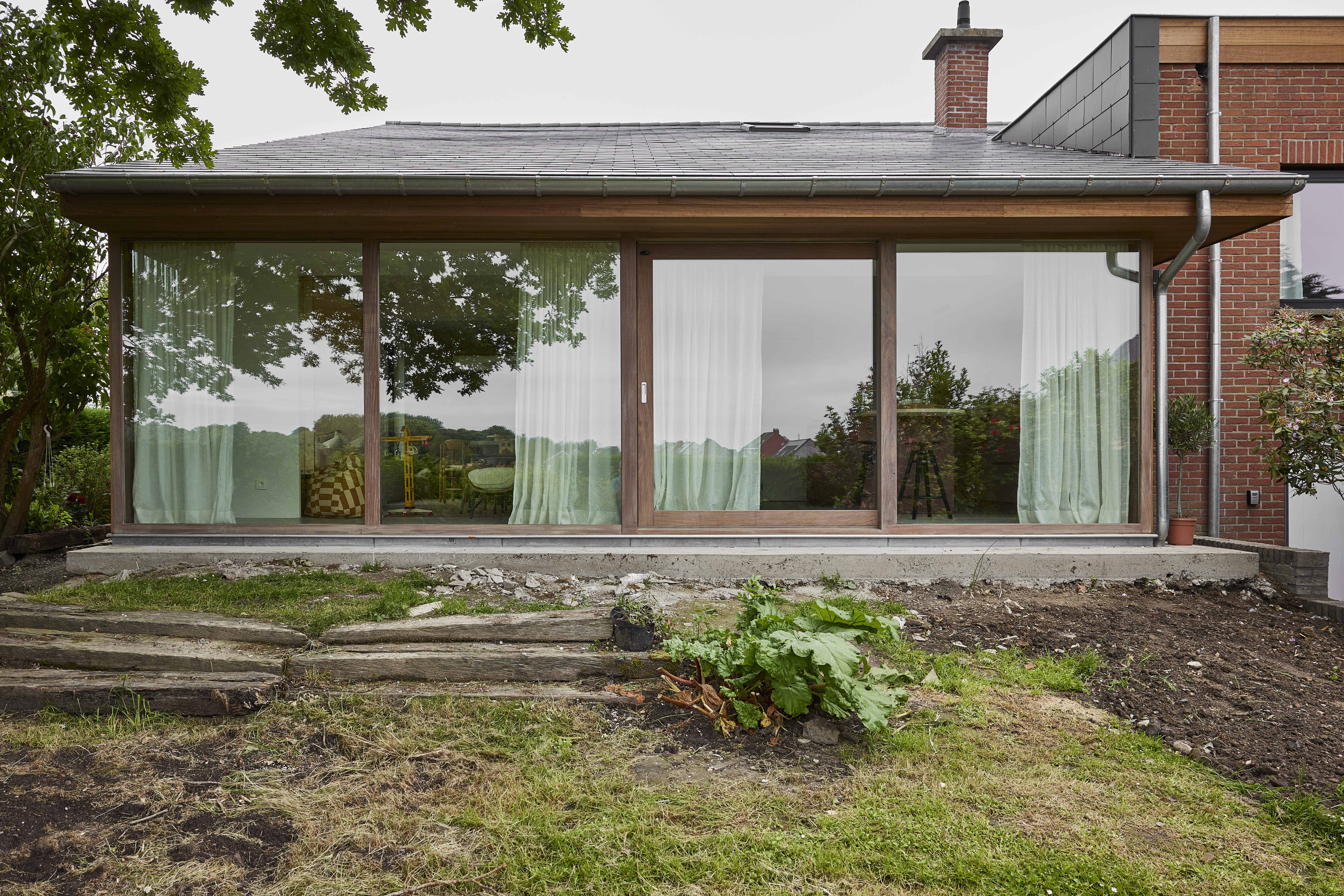 Grote glazen achtergevel met houten raamprofielen in Afromosia, behandeld met Rubio Natural, naast een bakstenen woning en een tuin met grond en gras.