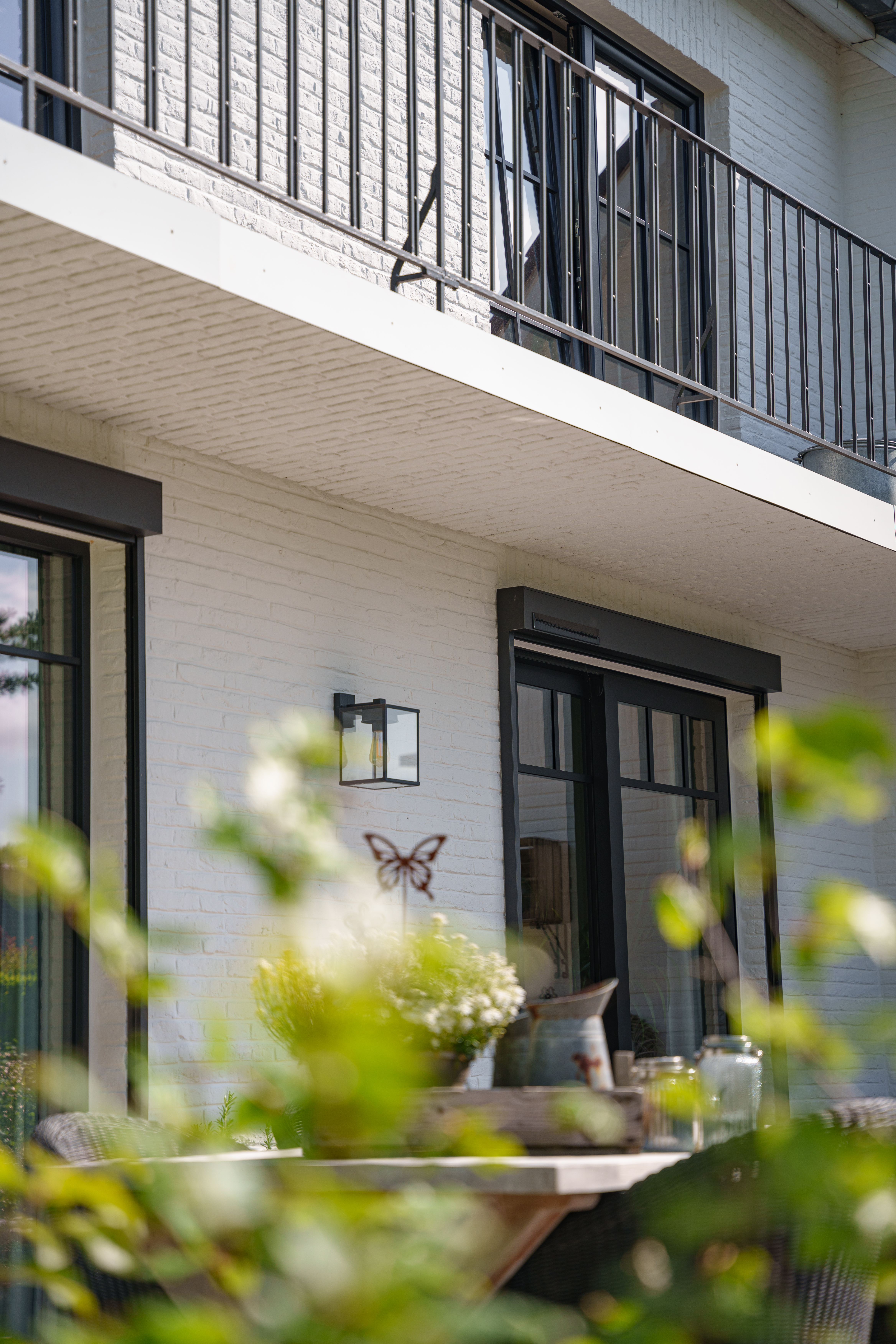 Witte woninggevel met zwarte balustrade, donkere raamprofielen en ronde zwarte voorzetrolluiken, met onscherpe beplanting op de voorgrond.