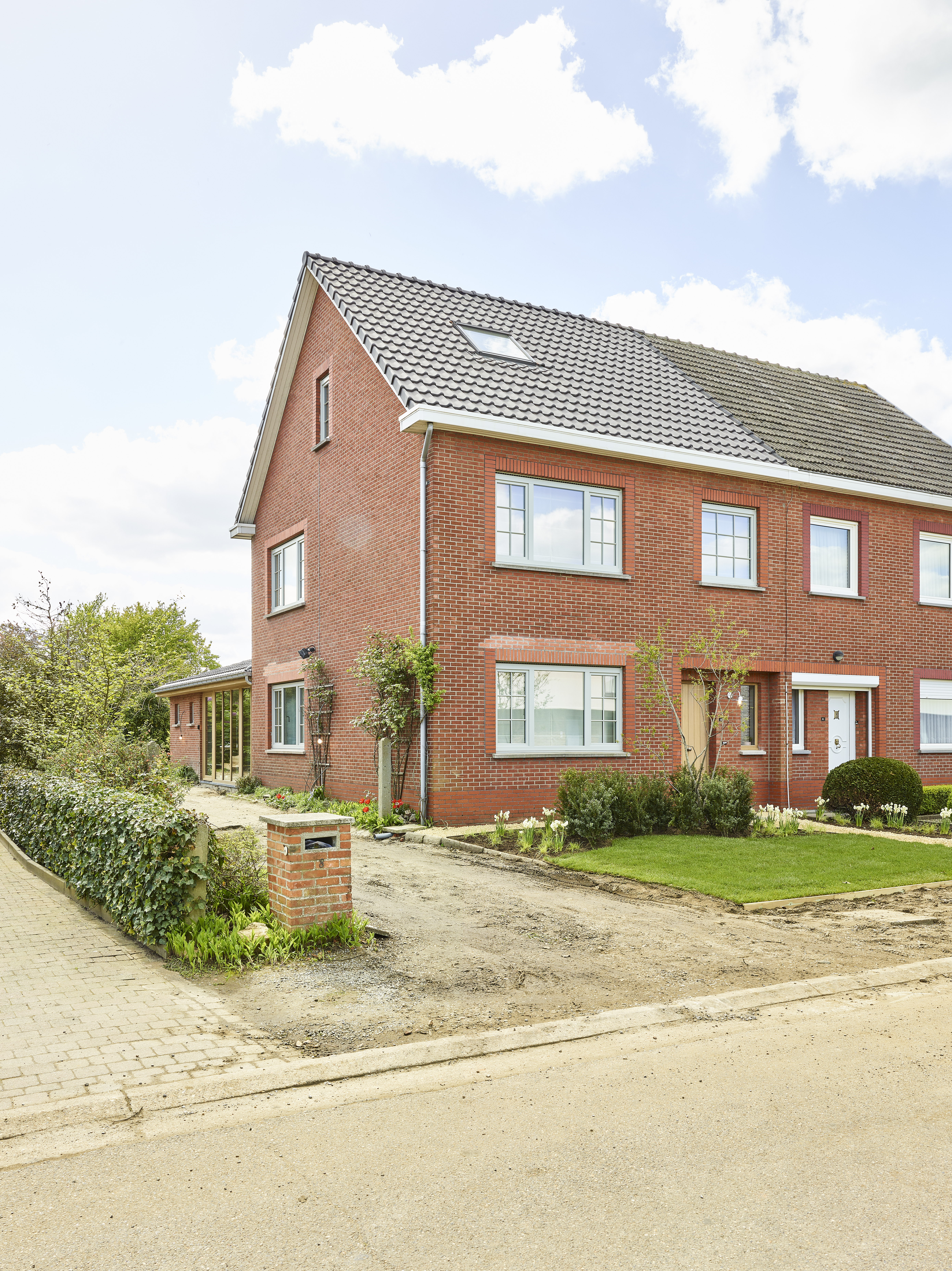 Rood bakstenen huis met lichte draai-kiepramen en driedubbele beglazing, omgeven door een voortuin en een verharde straat.