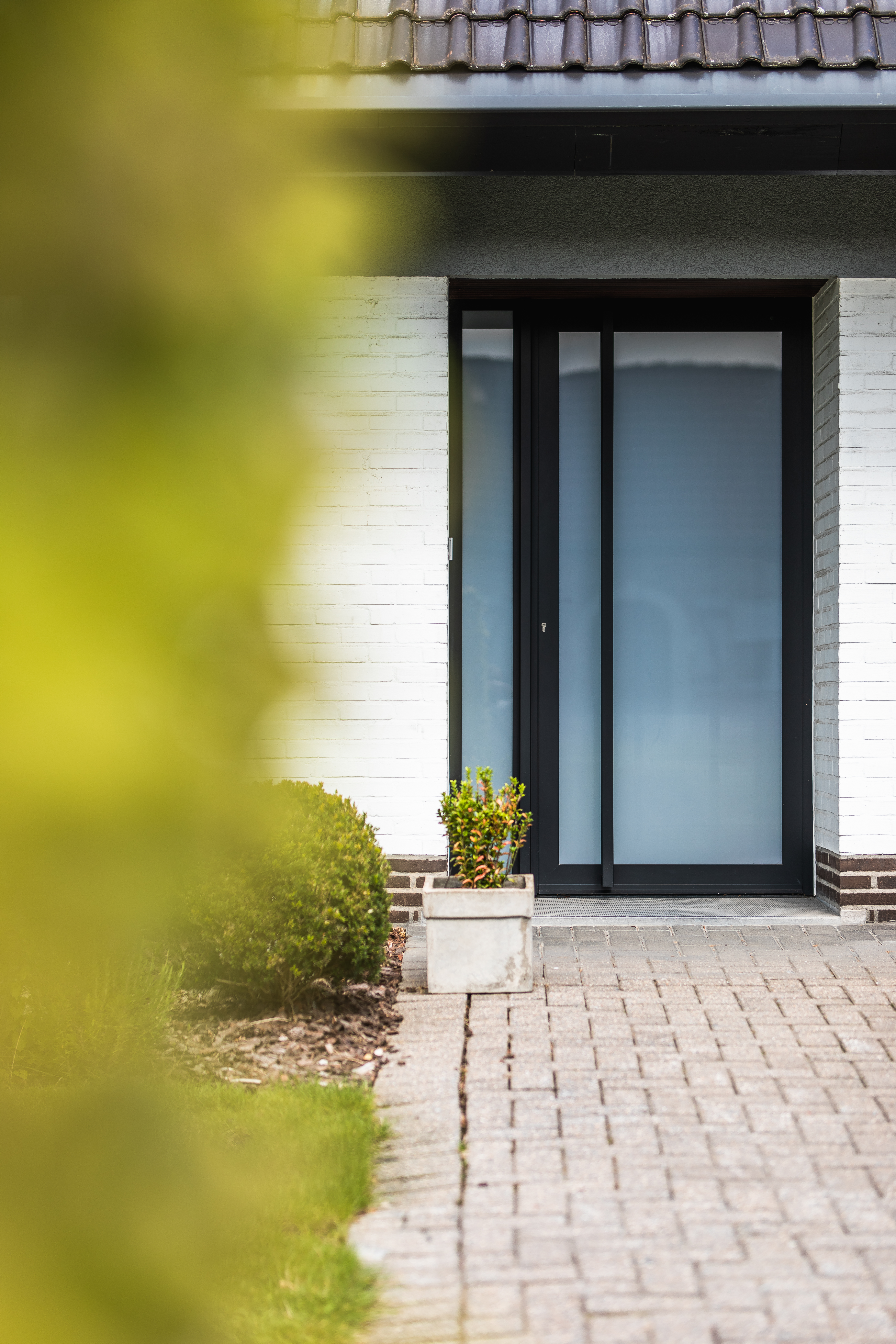 Moderne inkom van een open bebouwing met een zwarte beglaasde aluminiumdeur en smal zijlicht in een lichte bakstenen gevel.