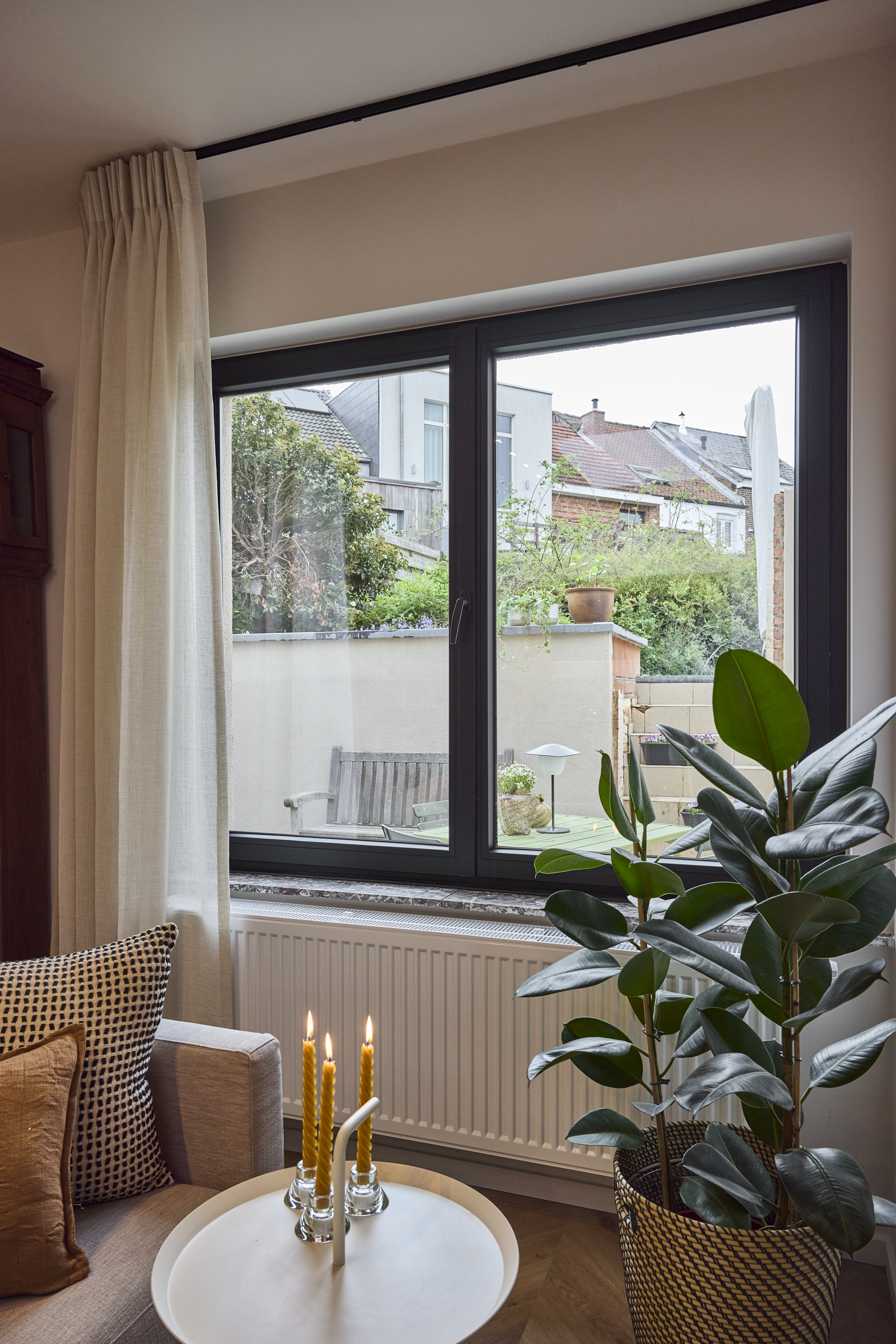 Zwart mat Komfort PLUS-dubbel raam met draai-kipfunctie in een lichte leefruimte, met uitzicht op een groene binnenkoer en omliggende woningen.