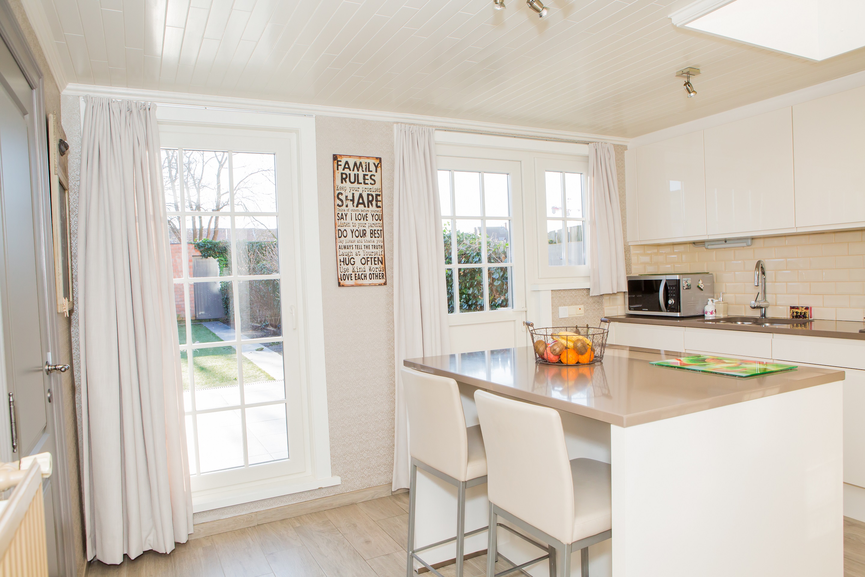Lichte, klassiek gerenoveerde keuken in een rijwoning met witte ramen en terrasdeur, kookeiland met zitplaatsen en accenten in licht hout.