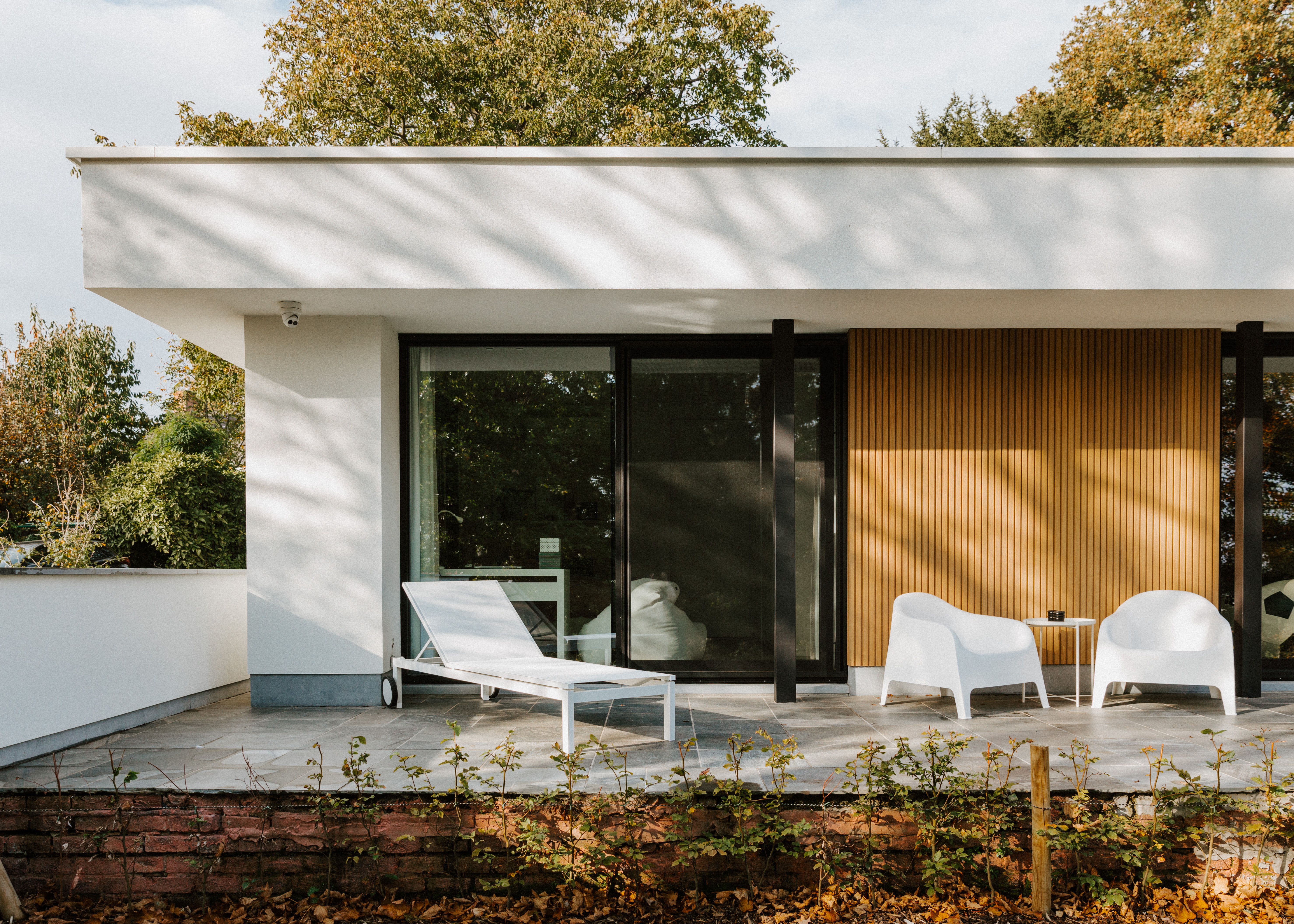 Moderne gerenoveerde vrijstaande woning met een zwart aluminium schuifraam en schuifvliegenraam, naast een verticale houten wandbekleding en terras met twee witte fauteuils.
