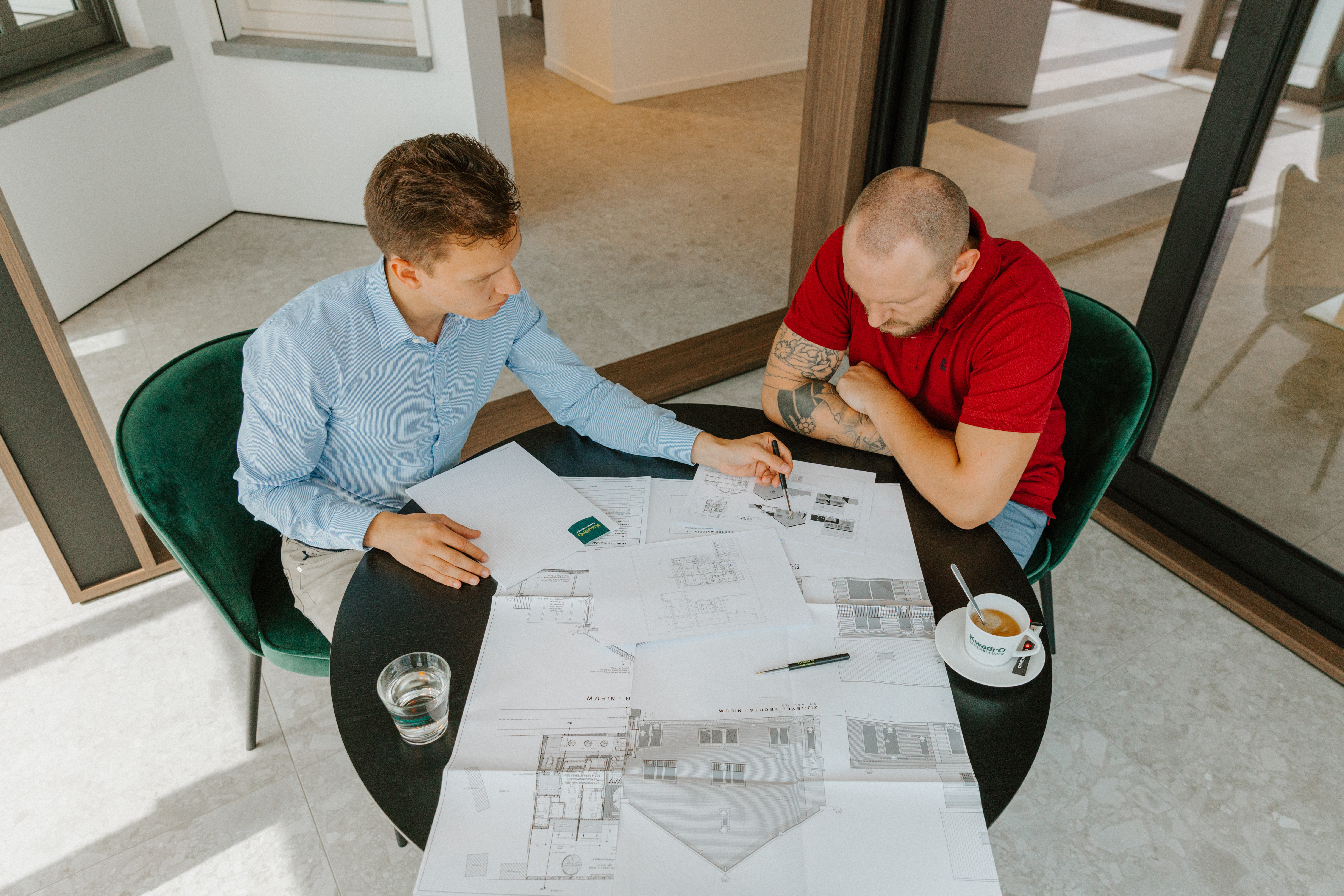 Twee mannen bespreken bouwplannen aan een tafel in een lichte ruimte naast grote zwarte schuiframen.