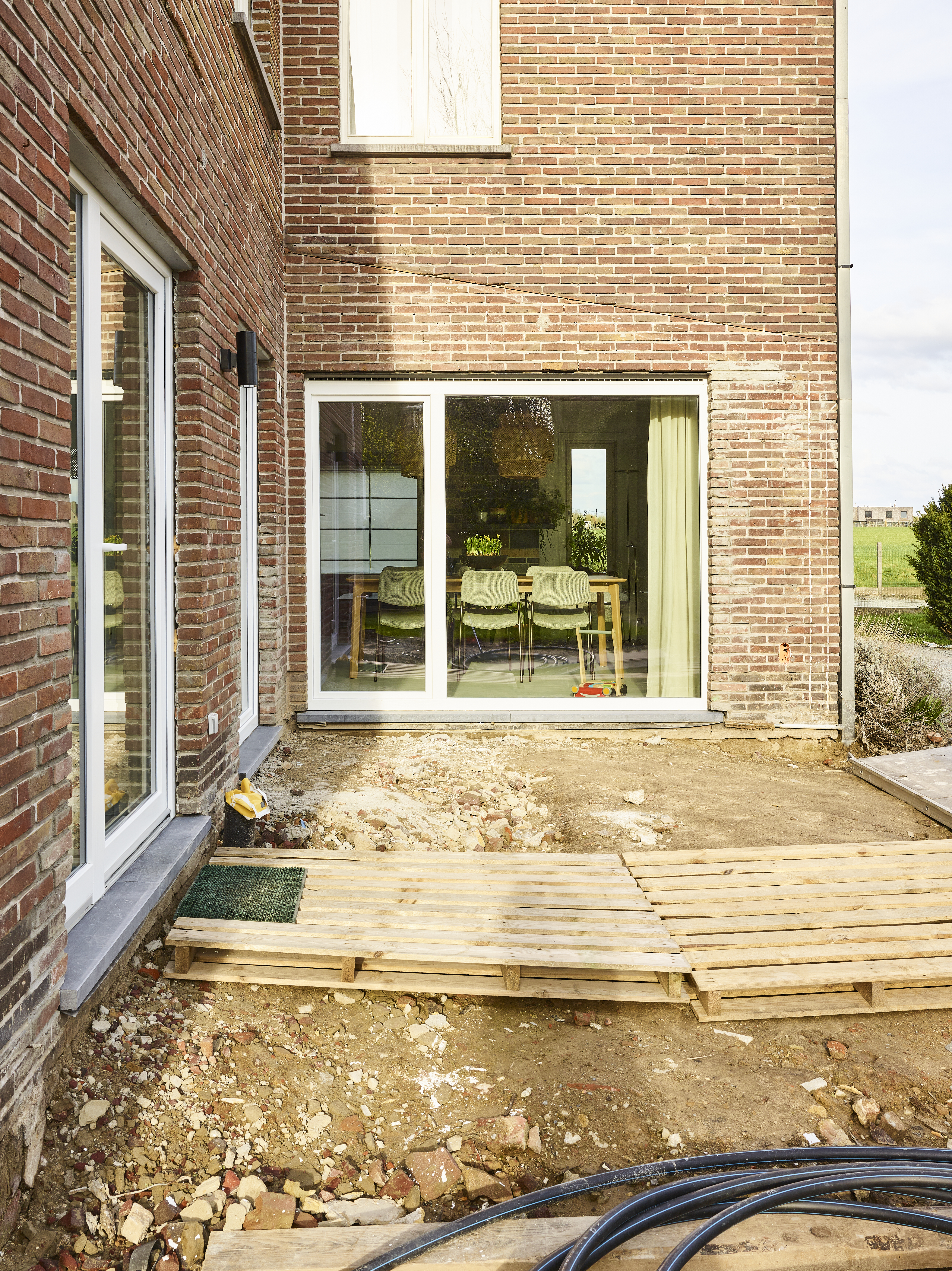 Wit draai-kipraam en vast raam in een gevel van rode baksteen, met zicht op de woonkamer en een nog onafgewerkt houten terras.