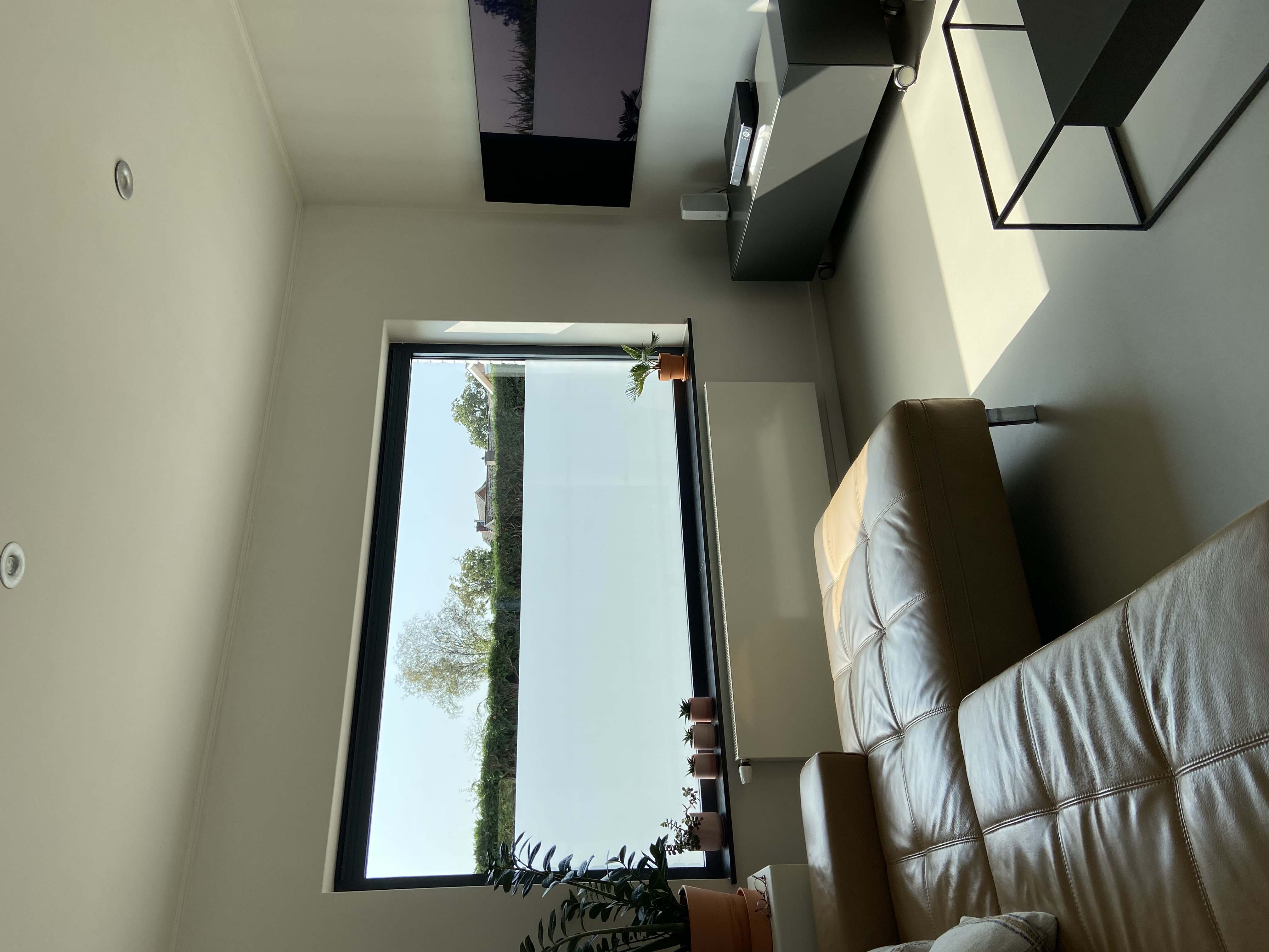 Moderne woonkamer van een vrijstaande woning met een groot raam met zwart profiel en zicht op het groen, een lederen zetel, salontafel en televisie aan de wand.