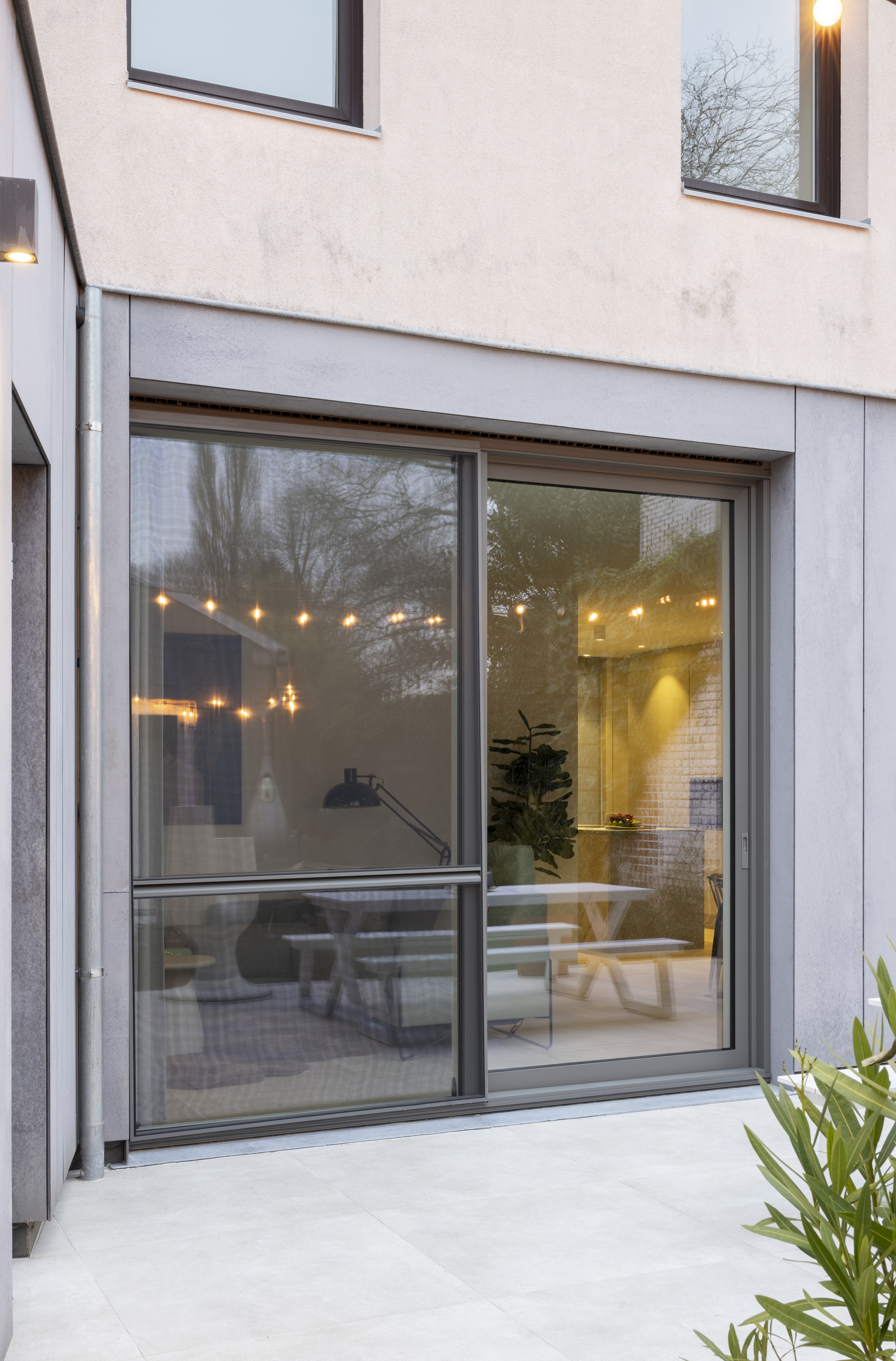 Moderne gerenoveerde gevel van een gesloten bebouwing met een gesloten grijs aluminium schuifraam en geïntegreerd schuifvliegenraam; binnen is een licht interieur zichtbaar.