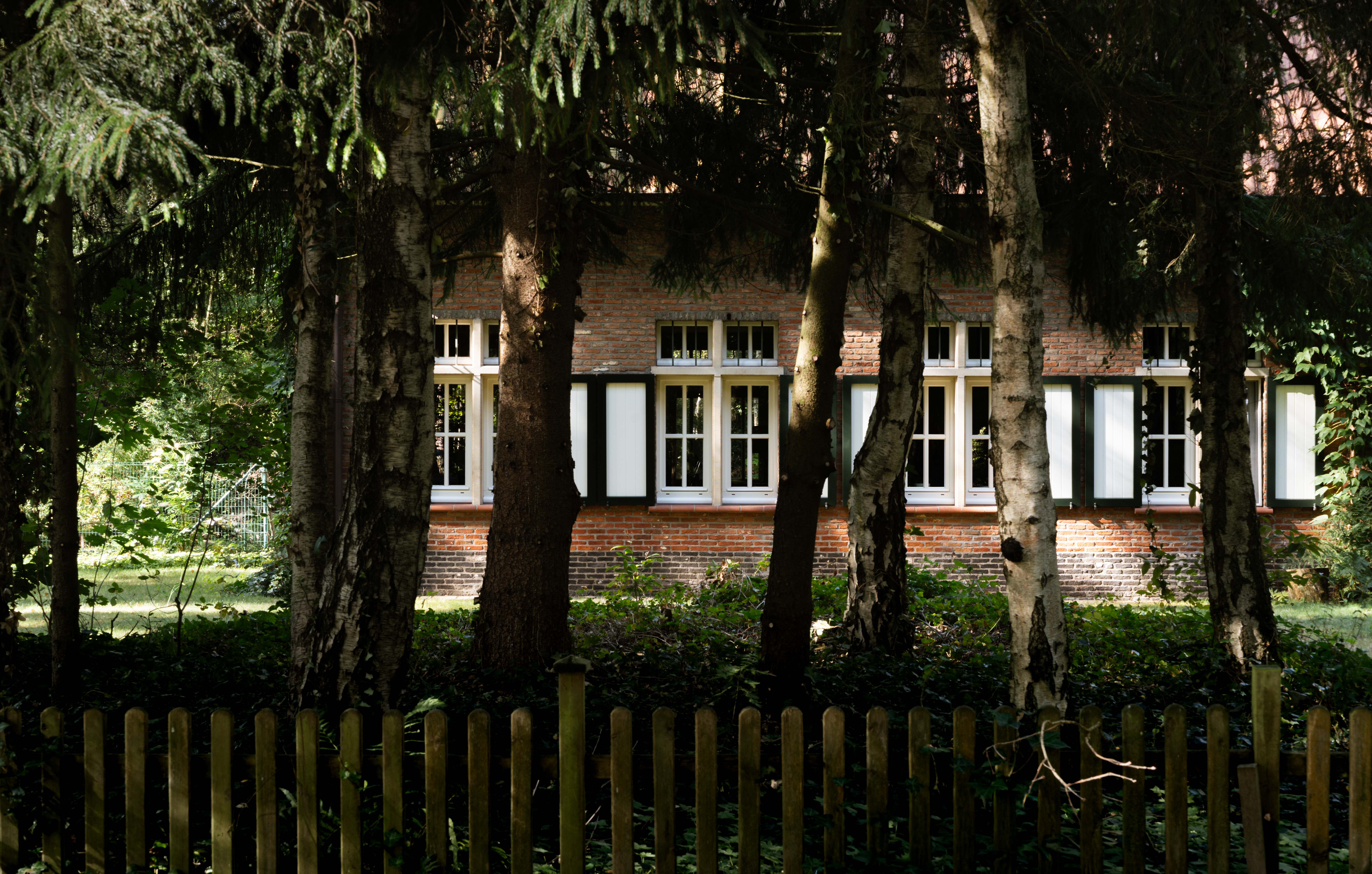 Een vrijstaande bakstenen woning in warme landelijke stijl, met lichte ramen die deels achter de bomen schuilgaan; de gerenoveerde gevel ligt in een groene omgeving.