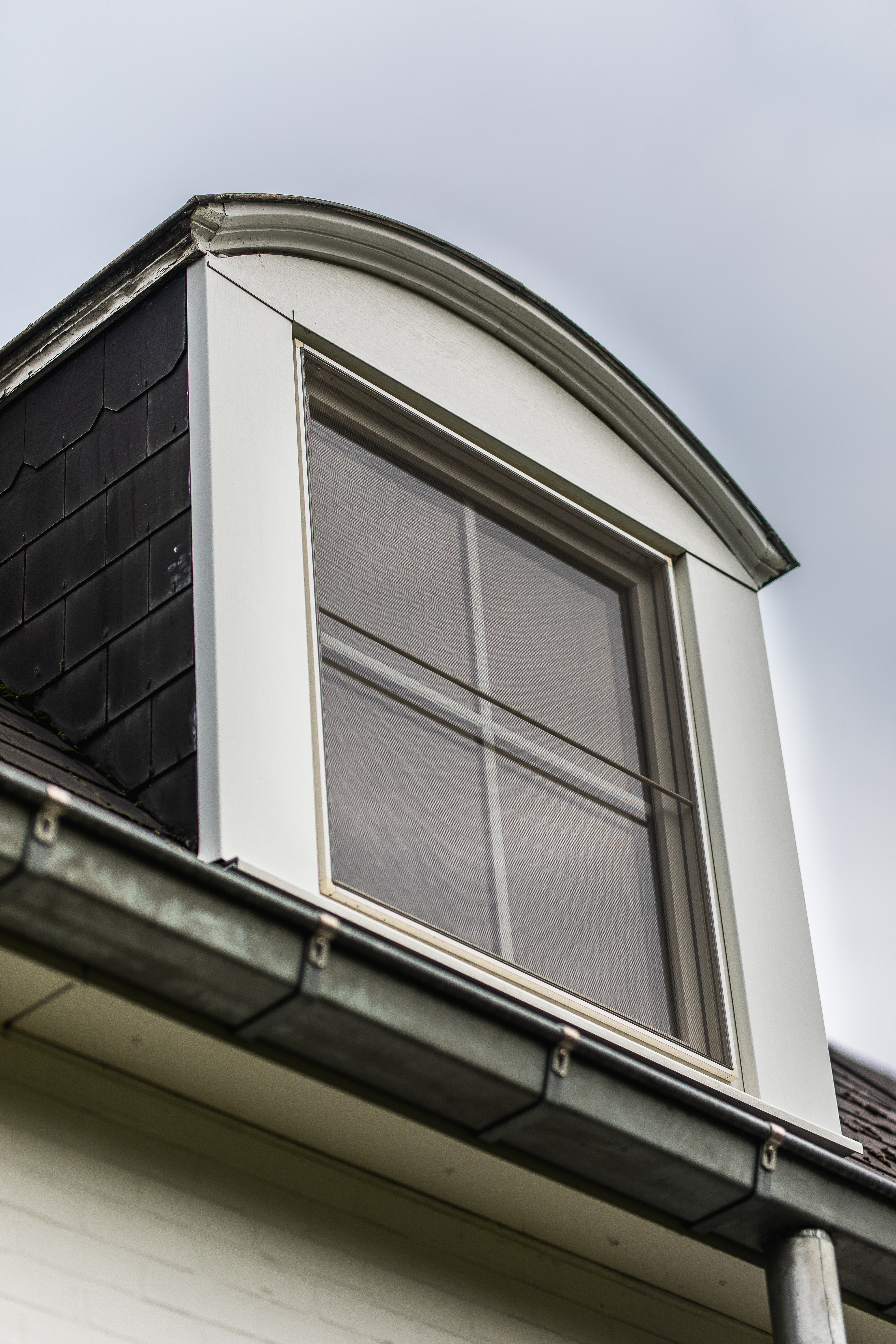 Klassiek vormgegeven raam in een witte dakkapel van een vrijstaande woning, verwerkt in een lichte gevel met donkere dakbekleding.