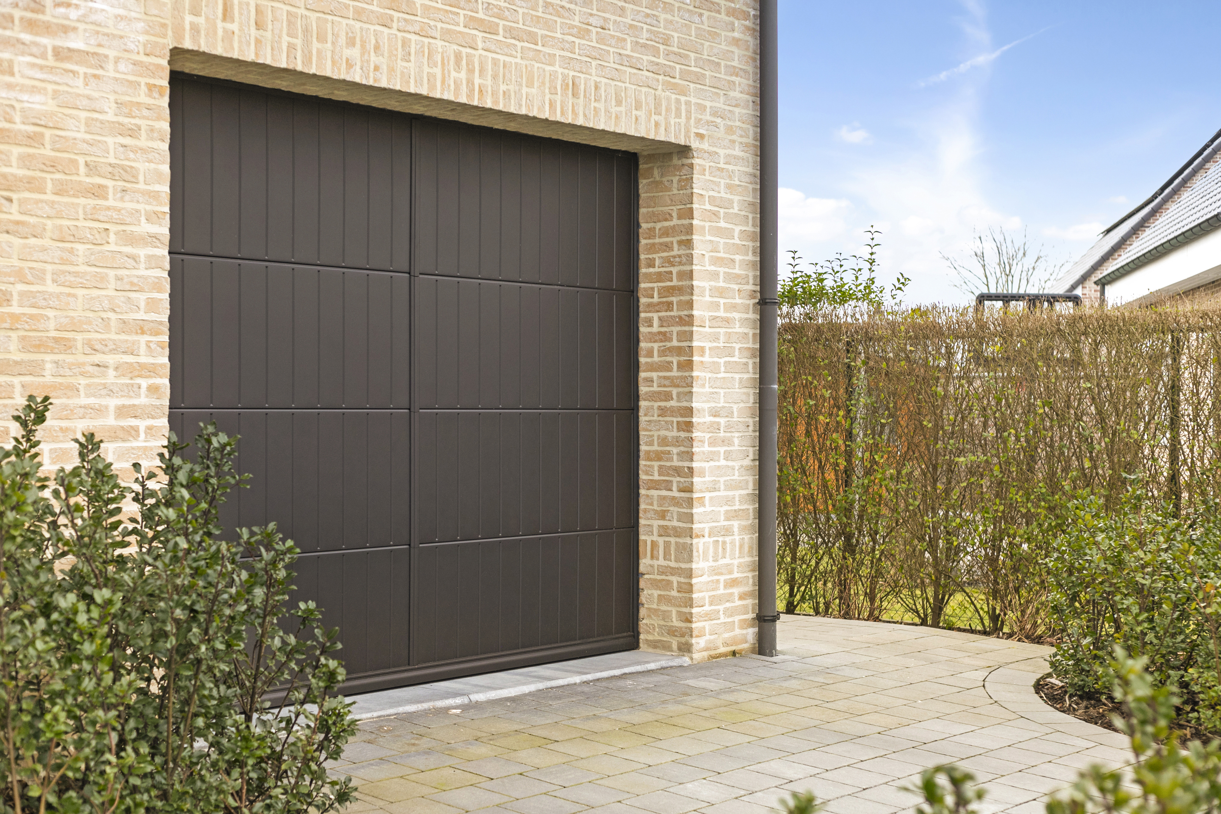 Zwart mat afgewerkte garagepoort in een warme landelijke bakstenen gevel, met betegelde oprit en haag bij een open bebouwing.