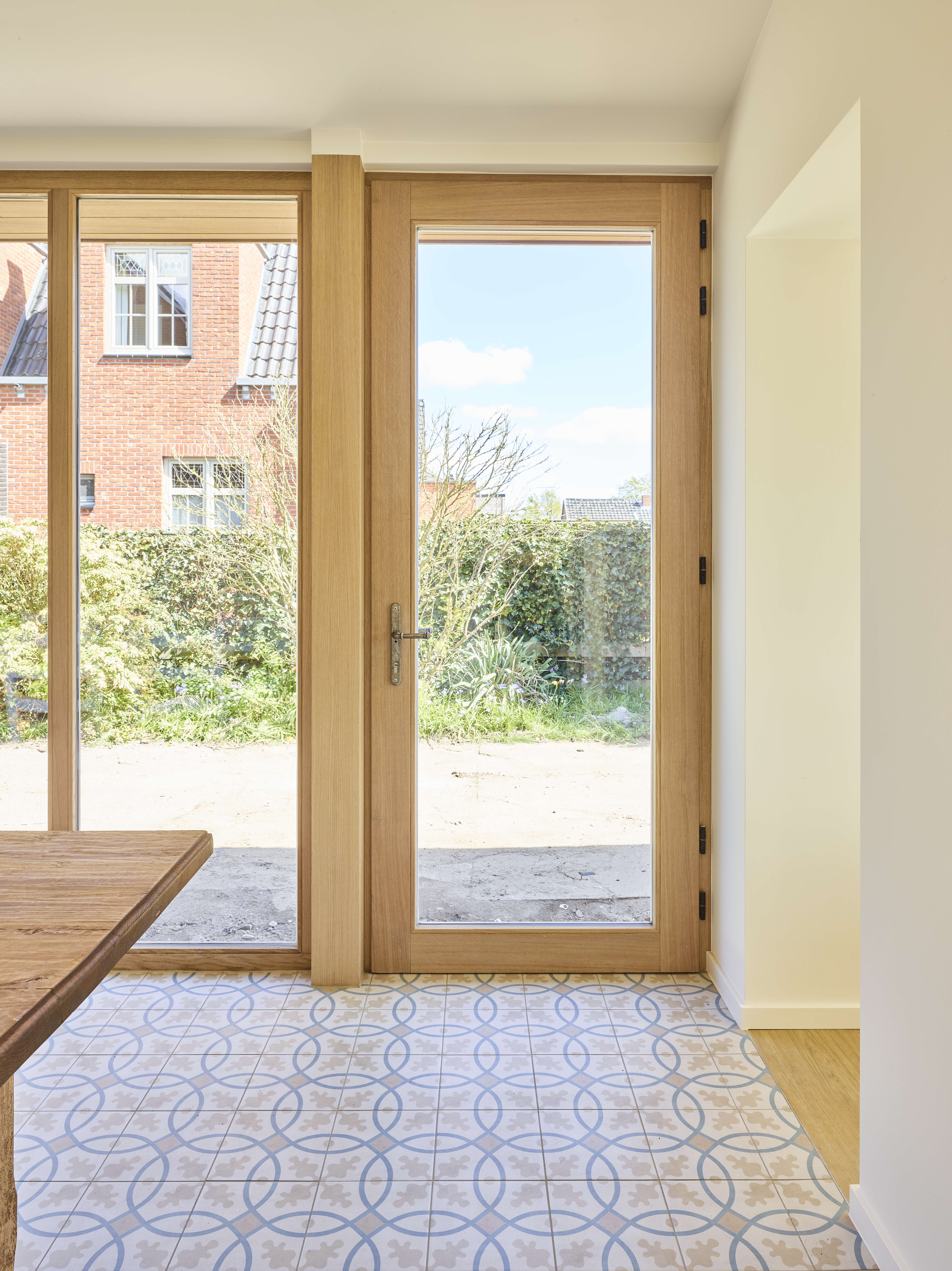 Beglaasde deur in gelakt licht eikenhout in een warm, landelijk interieur, naast een kamerhoog raam met uitzicht op de tuin.