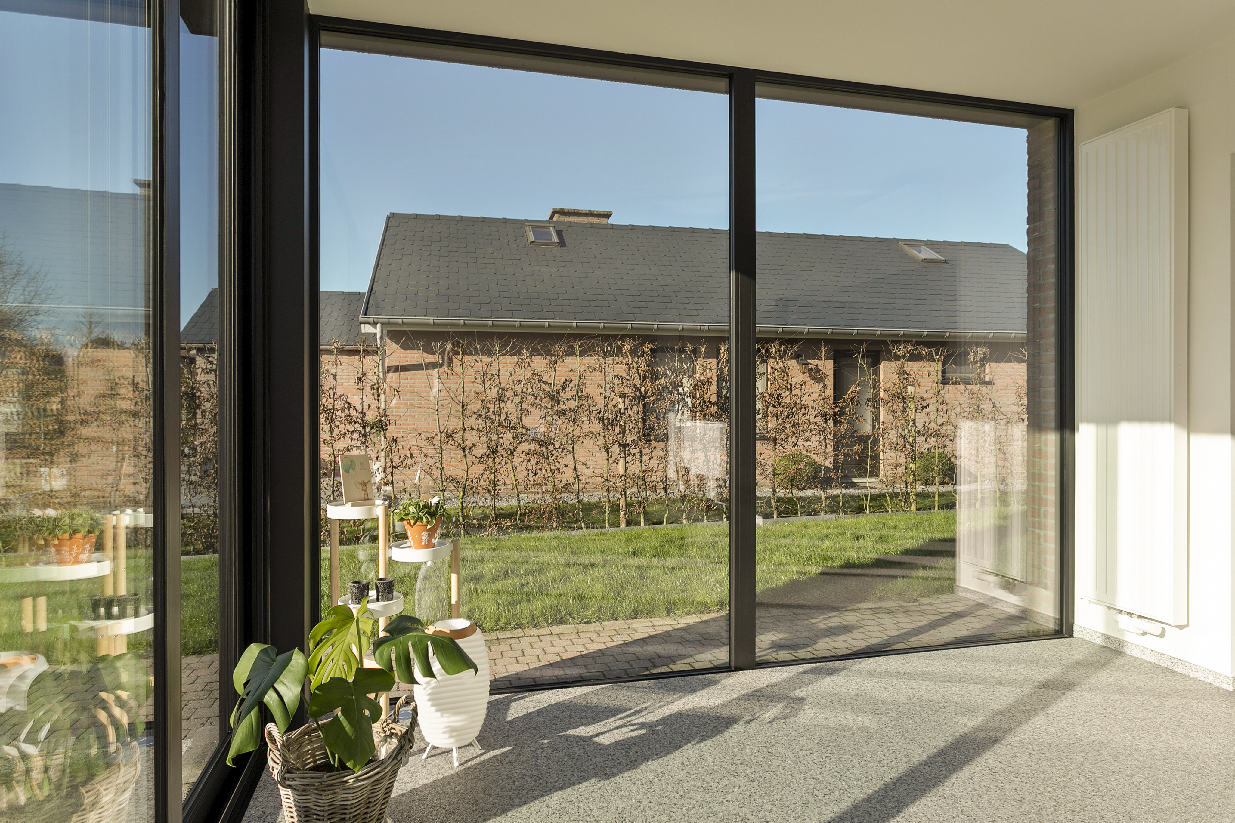 Groot vast raam met zwarte aluminiumprofielen in een gerenoveerde leefruimte, met uitzicht op een tuin en een tegenoverliggende woning.