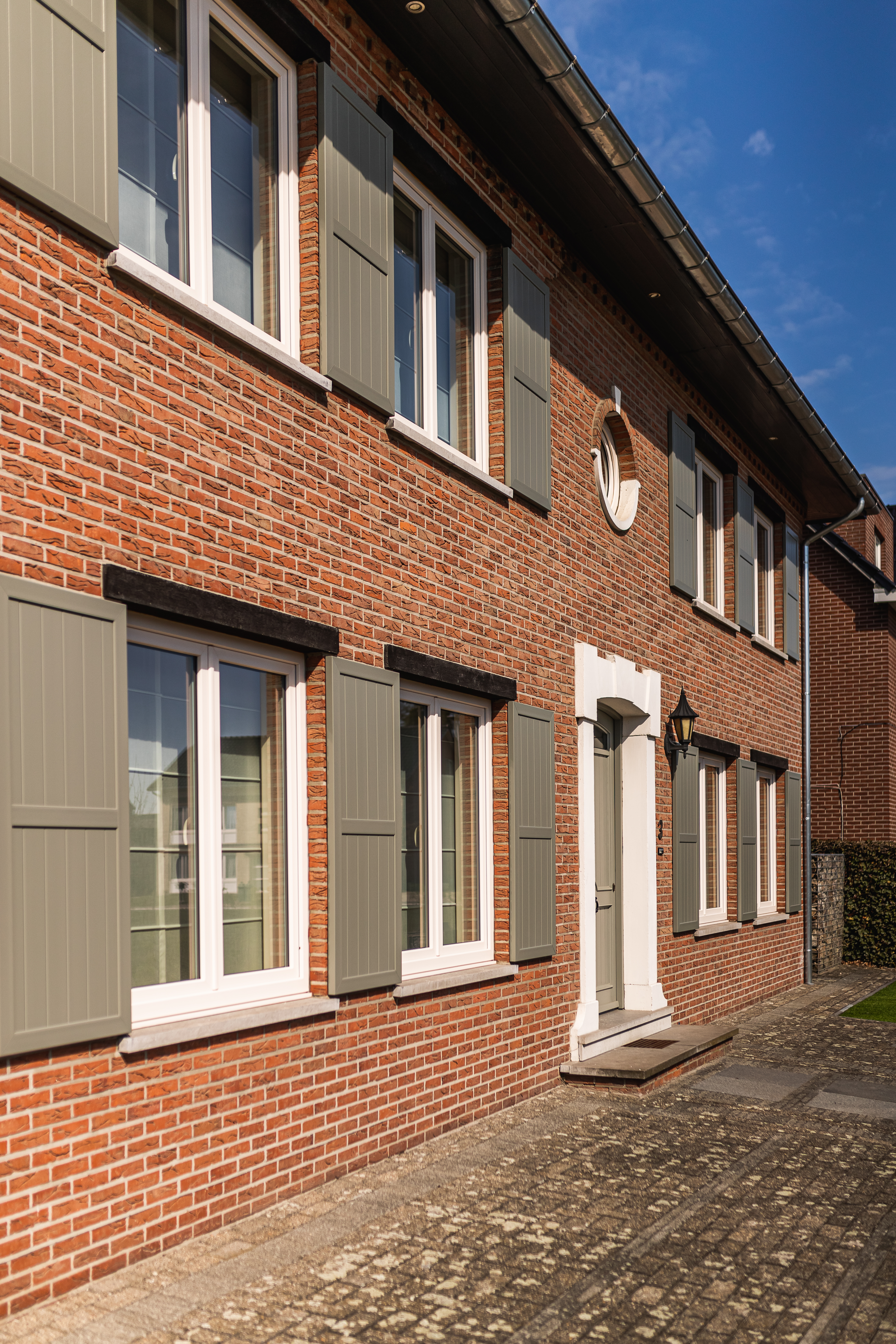 Gerenoveerde bakstenen gevel van een vrijstaande woning met dubbele ramen in crème NERF-afwerking, grijze klapluiken en een inkomdeur langs de verharde oprit.