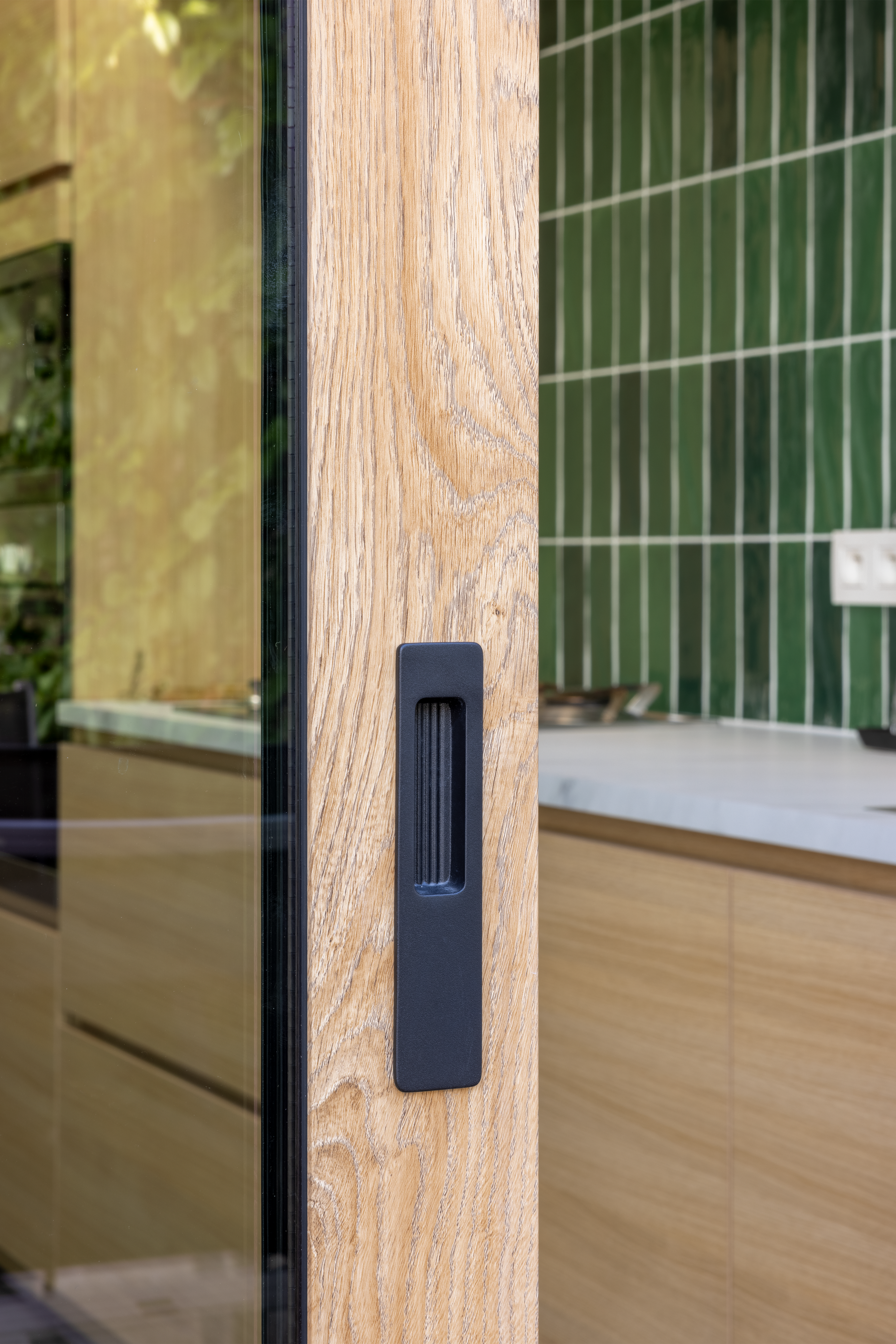 Close-up van een schuifraam met Woodec Eik Natuur NERF-houtdecor en zwarte handgreep, in een keuken met groene tegels en houten fronten.