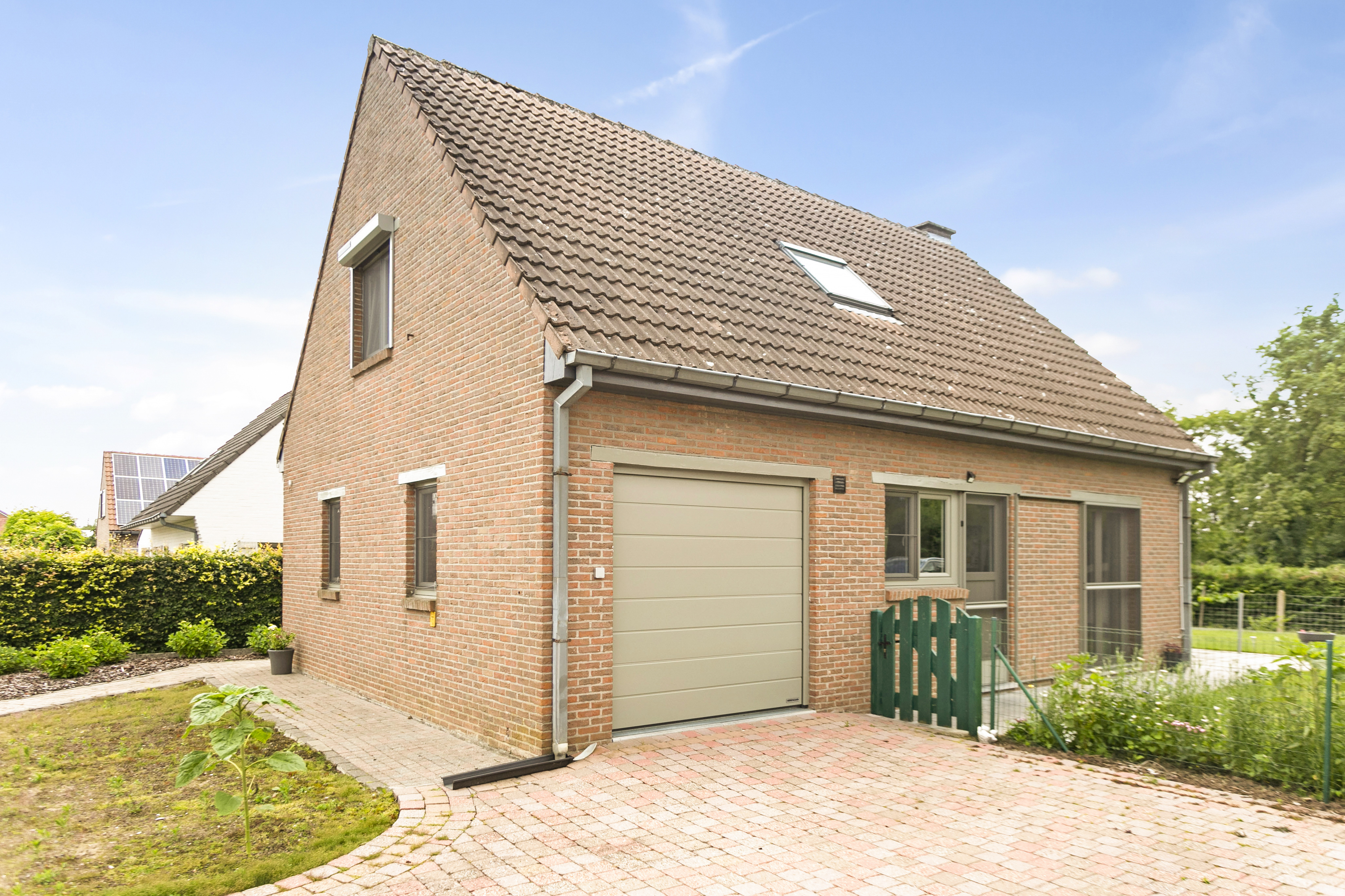 Open bebouwing in baksteen met een betongrijze garagepoort, draai-kipramen en dubbele ramen, gezien vanaf de verharde oprit.