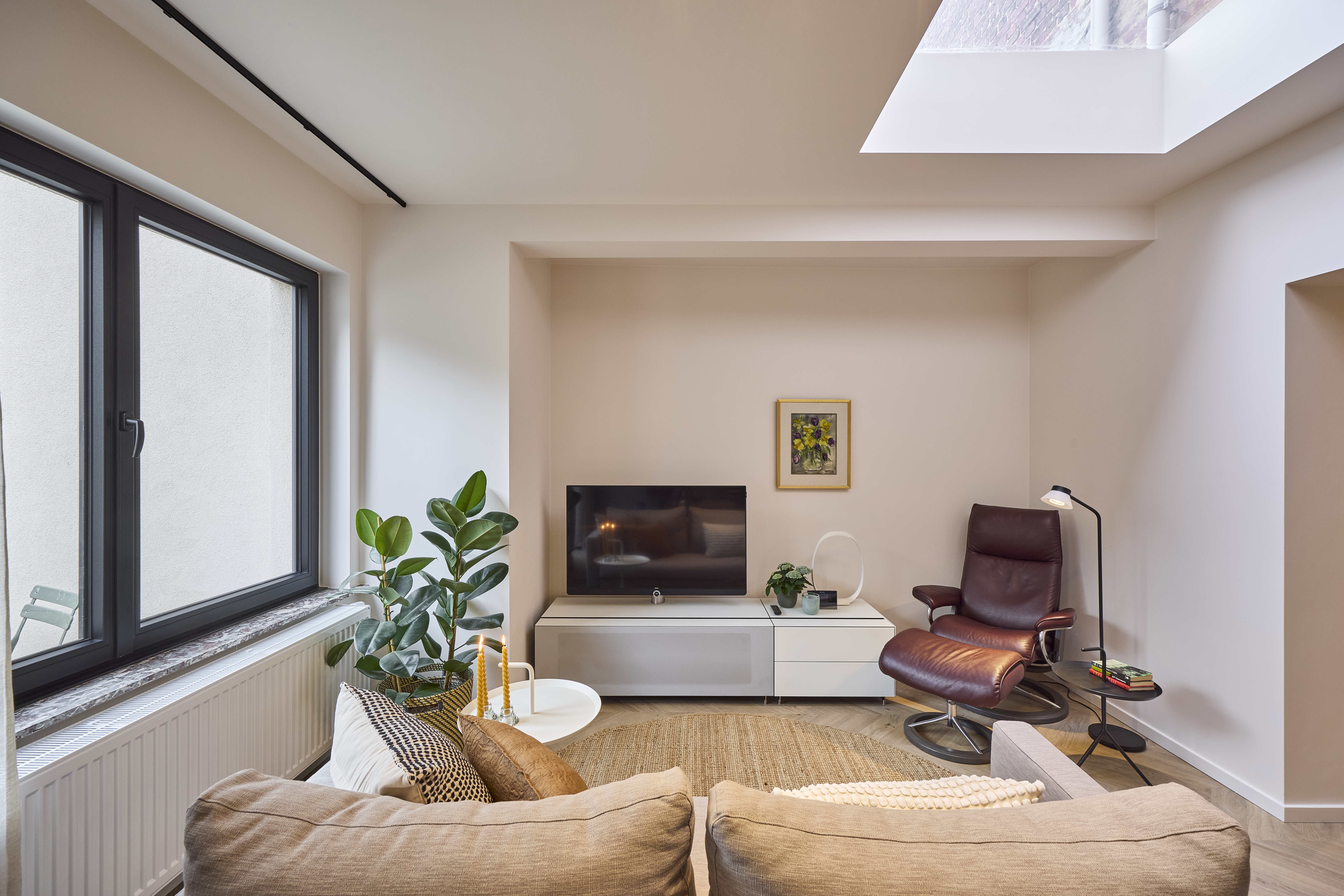 Lichte woonkamer met matzwart dubbel draai-kipraam, lederen fauteuil, sofa, televisie en dakraam.