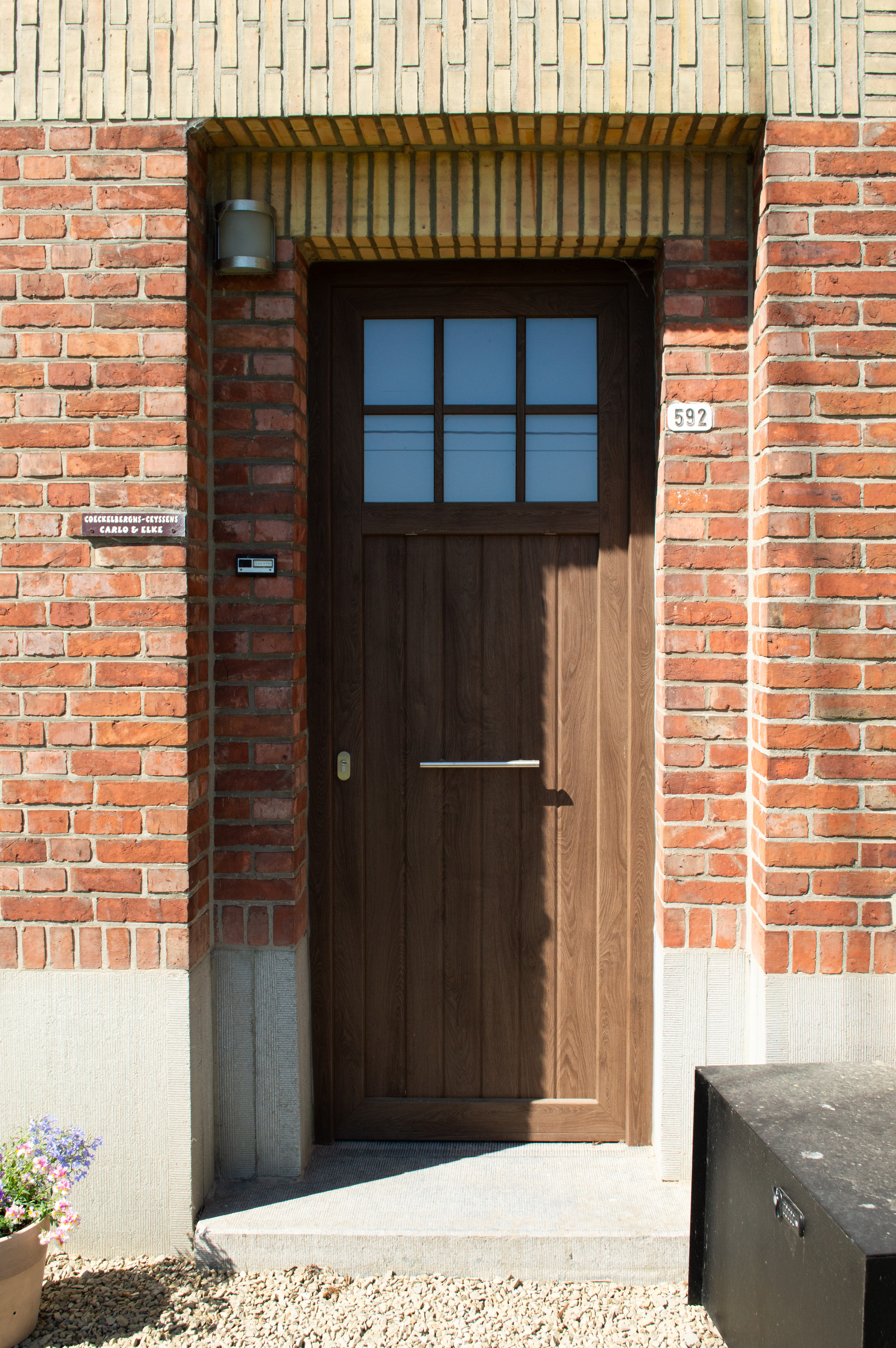 Donker houtlook-PVC-buitendeur met Sablé-afwerking en gedeeltelijke beglazing, geplaatst in een rode bakstenen gevel onder een gemetselde luifel.