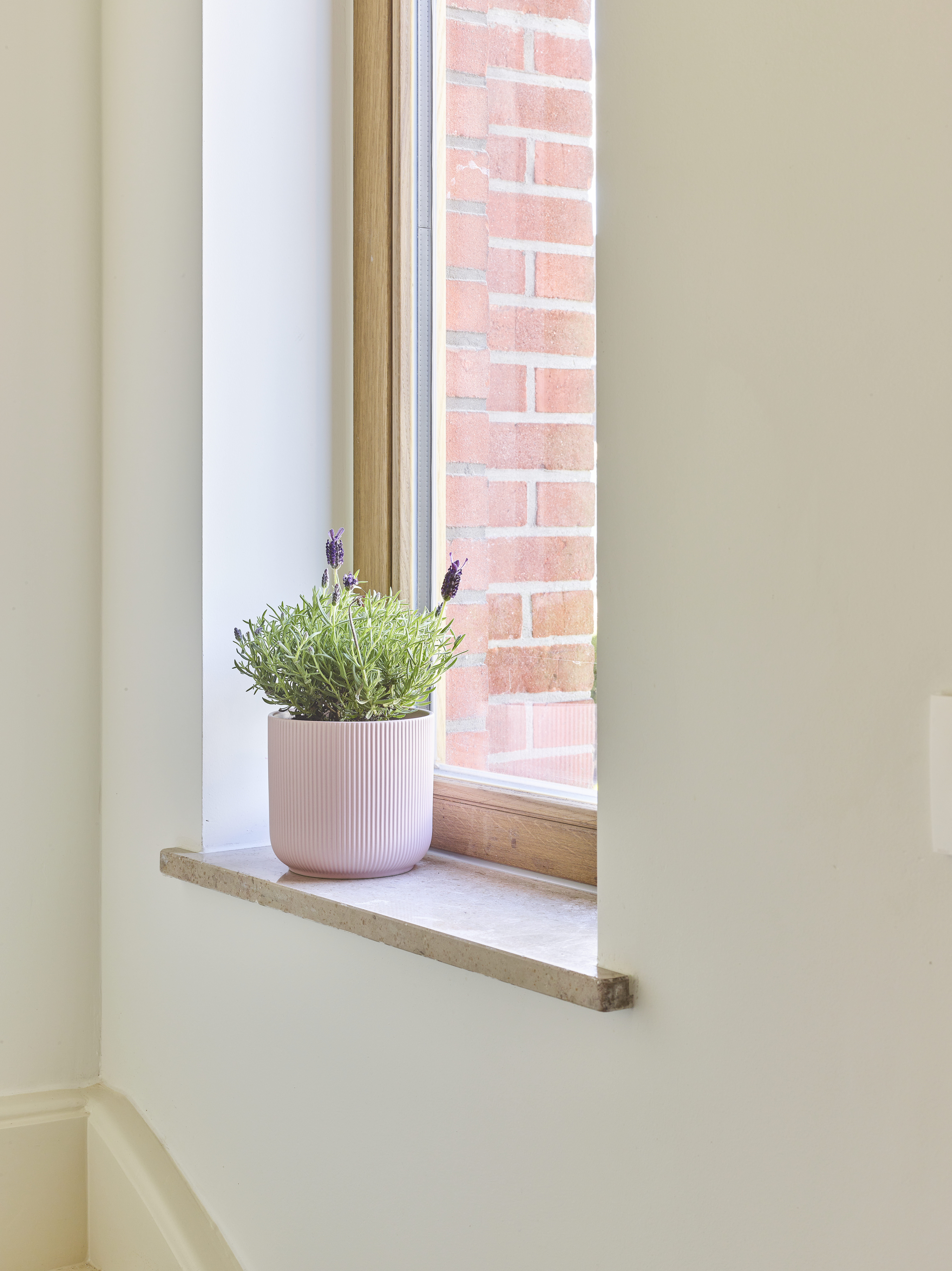 Vast raam met afgelakt raamprofiel in lichte eik, witte vensterbank en plant in pot, met zicht op een rode bakstenen gevel in een warme, landelijke inrichting.
