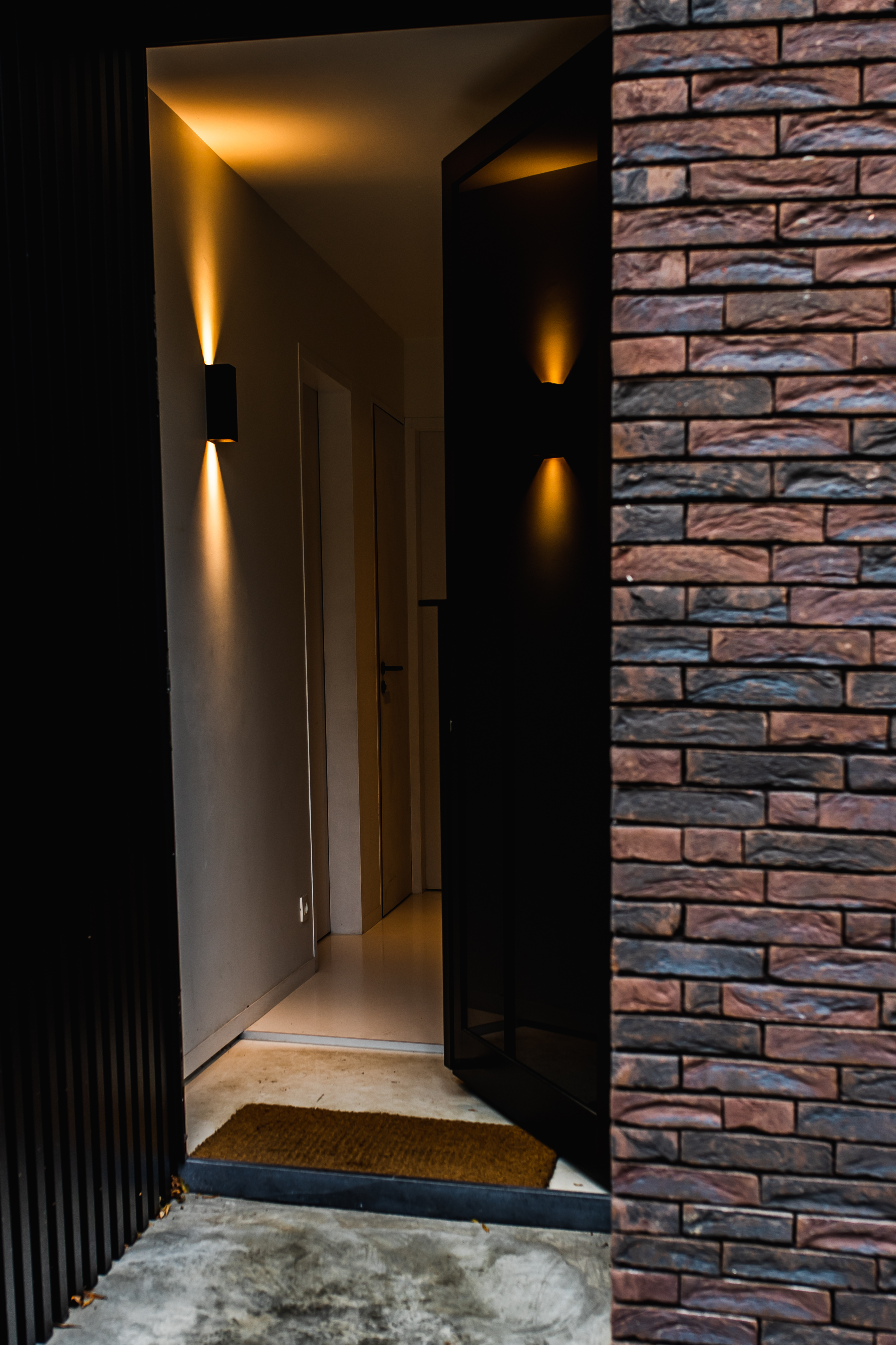 Moderne inkom van een nieuwbouwwoning met zwarte deur in 9005TC, karaktervolle bakstenen gevel en warm verlichte hal.