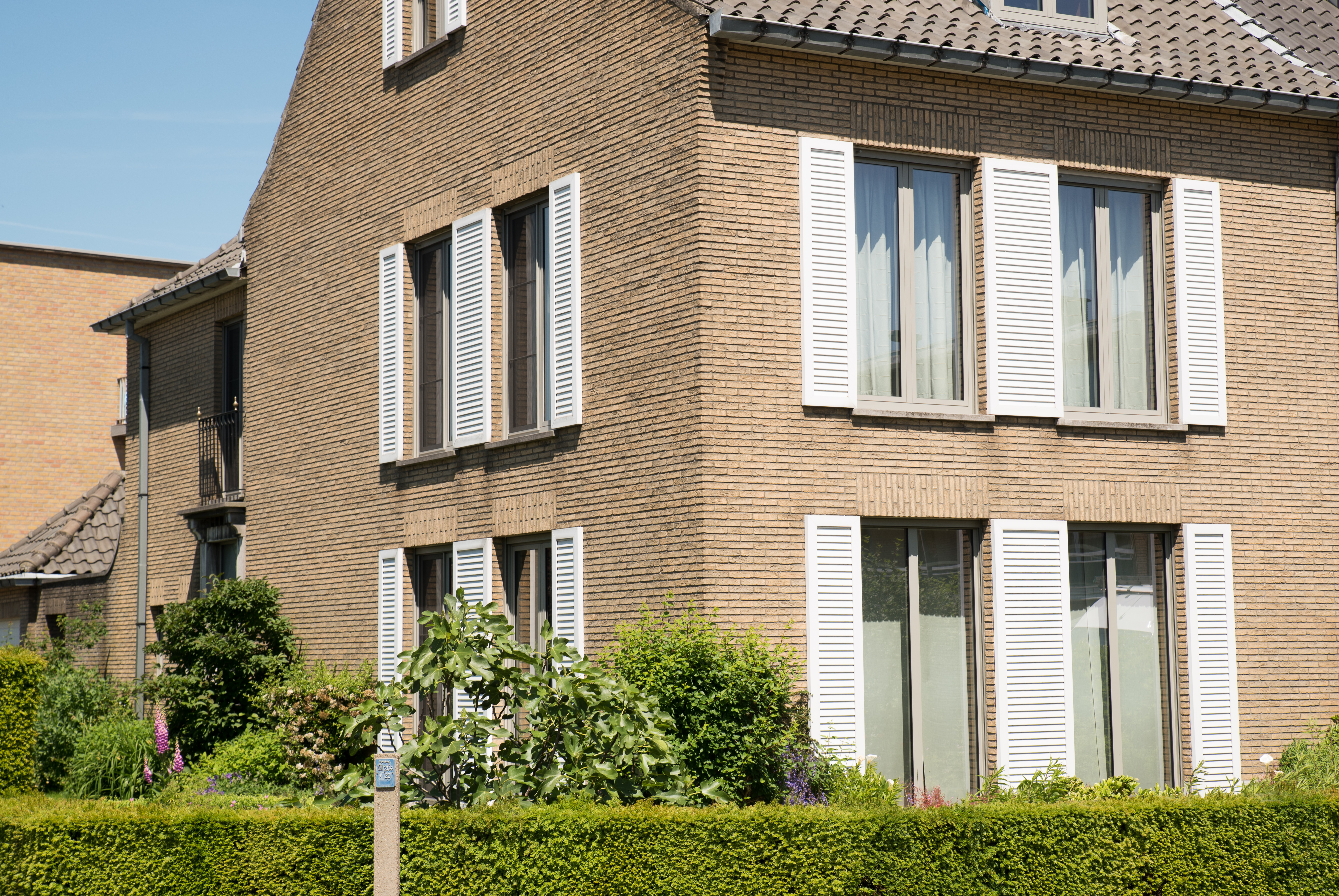 Renovatie van een driegevelwoning met bruine bakstenen gevel, witte luiken en grote ramen, zichtbaar boven een verzorgde voortuin.