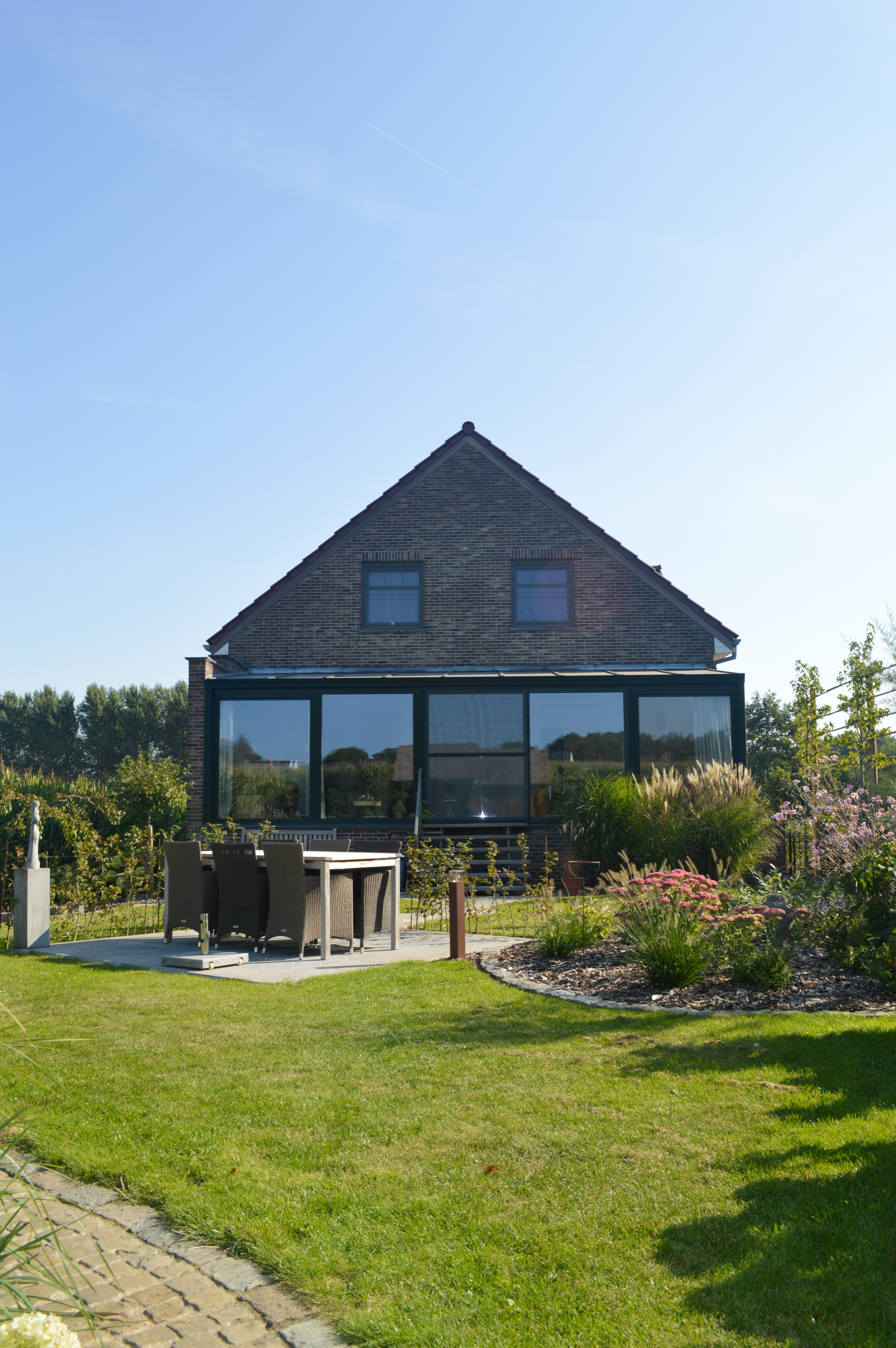 Achteraanzicht van een open bebouwing met overdekte glazen veranda, kwartsgrijze raamprofielen en verzorgde tuin; een schuifvliegenraam maakt de renovatie compleet.