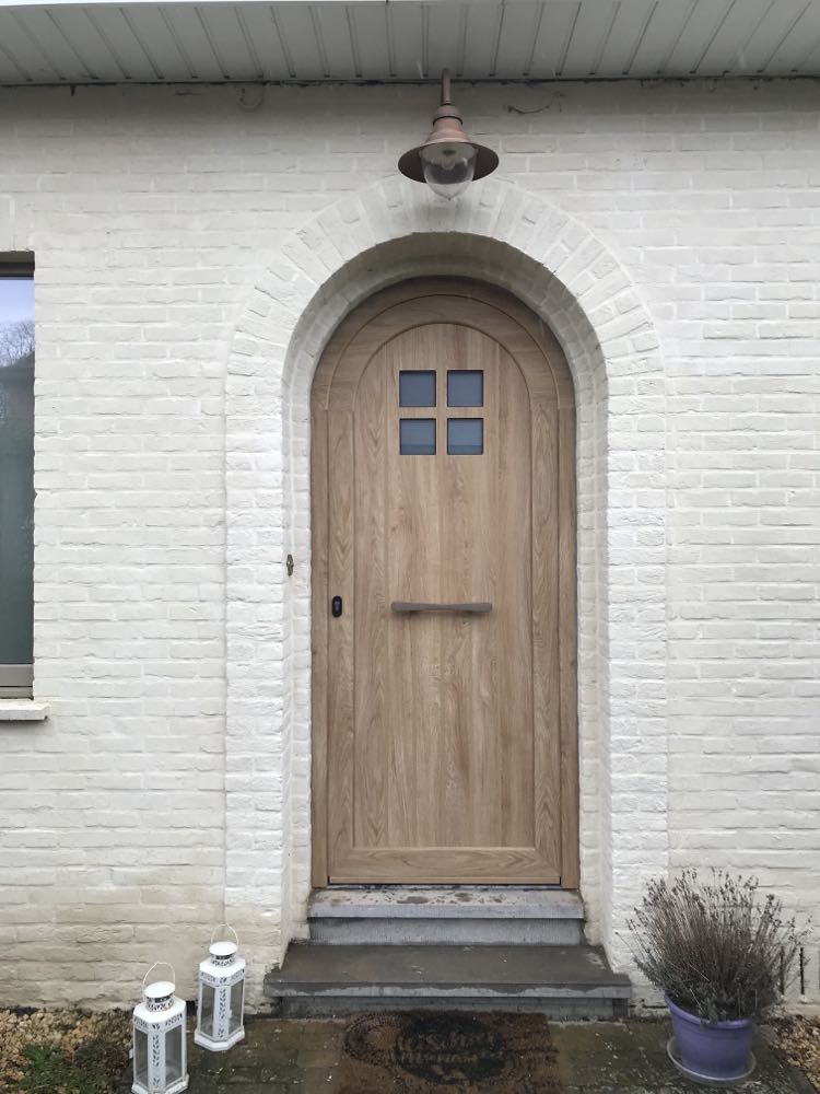 Panneau de porte en PVC cintré, finition Woodec Chêne Nature NERF, avec quatre petites fenêtres, intégré dans une façade en briques blanches.