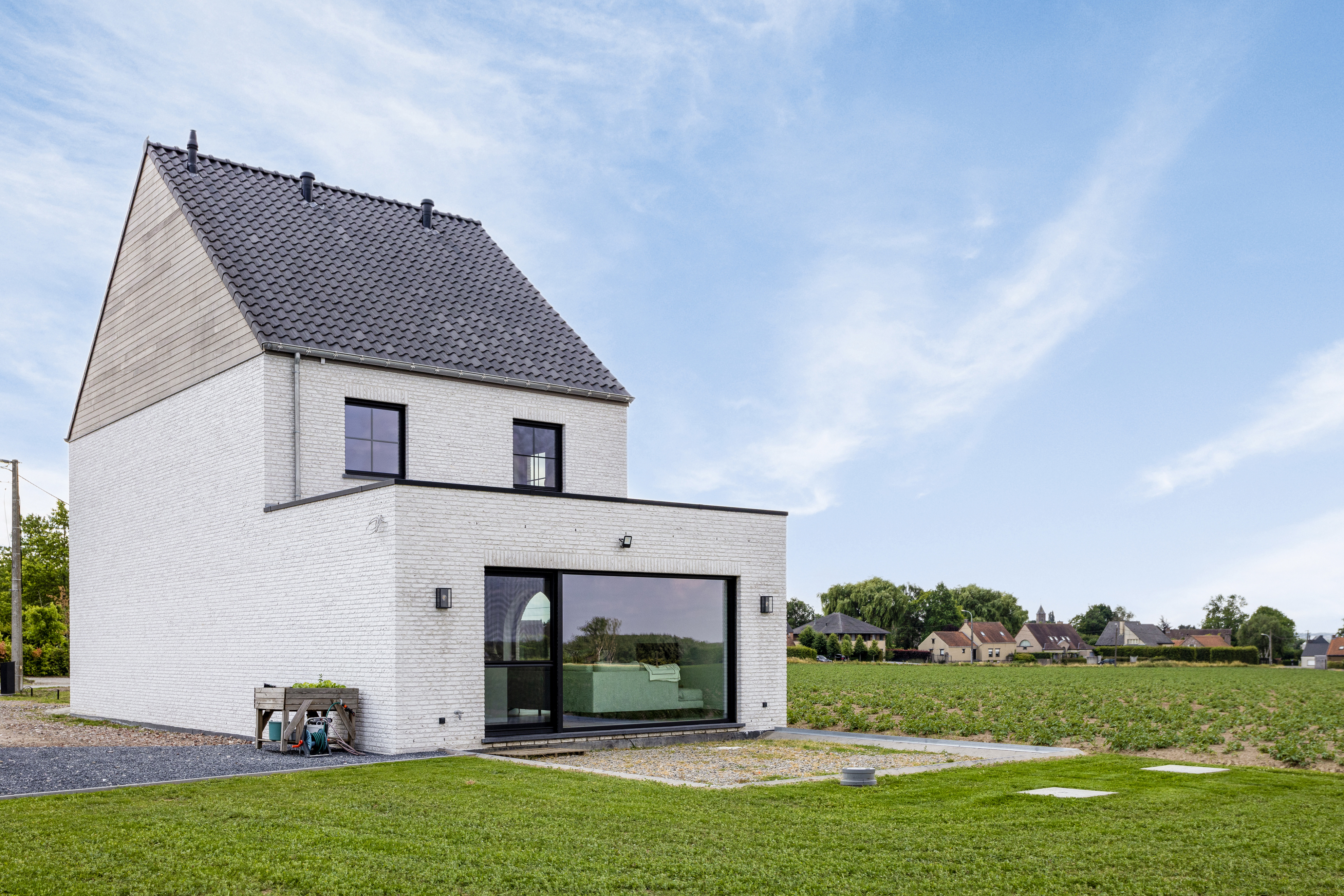 Moderne nieuwbouwwoning in open bebouwing met een groot vast raam in signaalzwart, geïntegreerd in een witte bakstenen gevel in een landelijke omgeving.