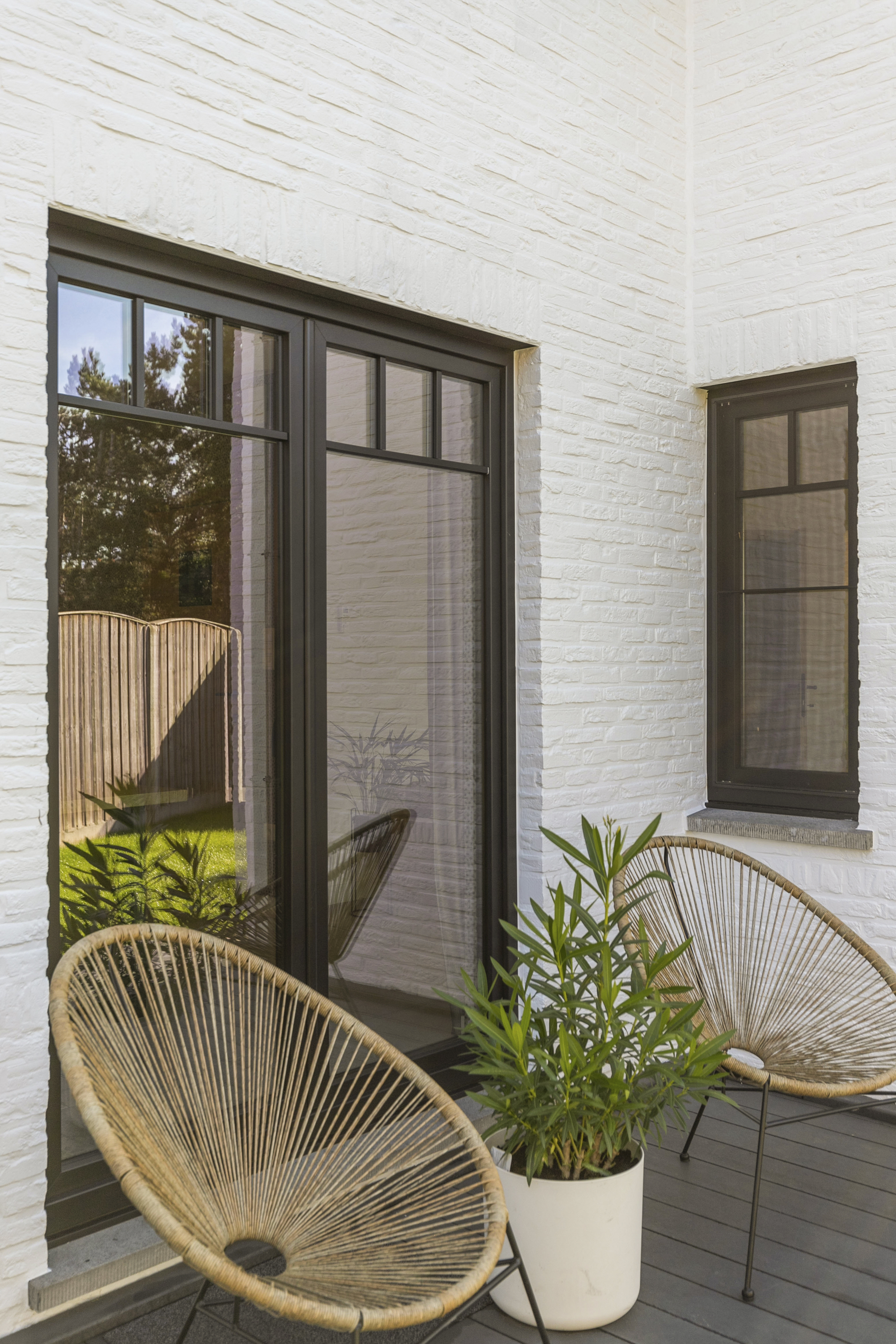 Mat zwart draai-kipraam en dubbel raam in een gerenoveerde lichte bakstenen gevel, met planten en rotanstoelen op het terras.