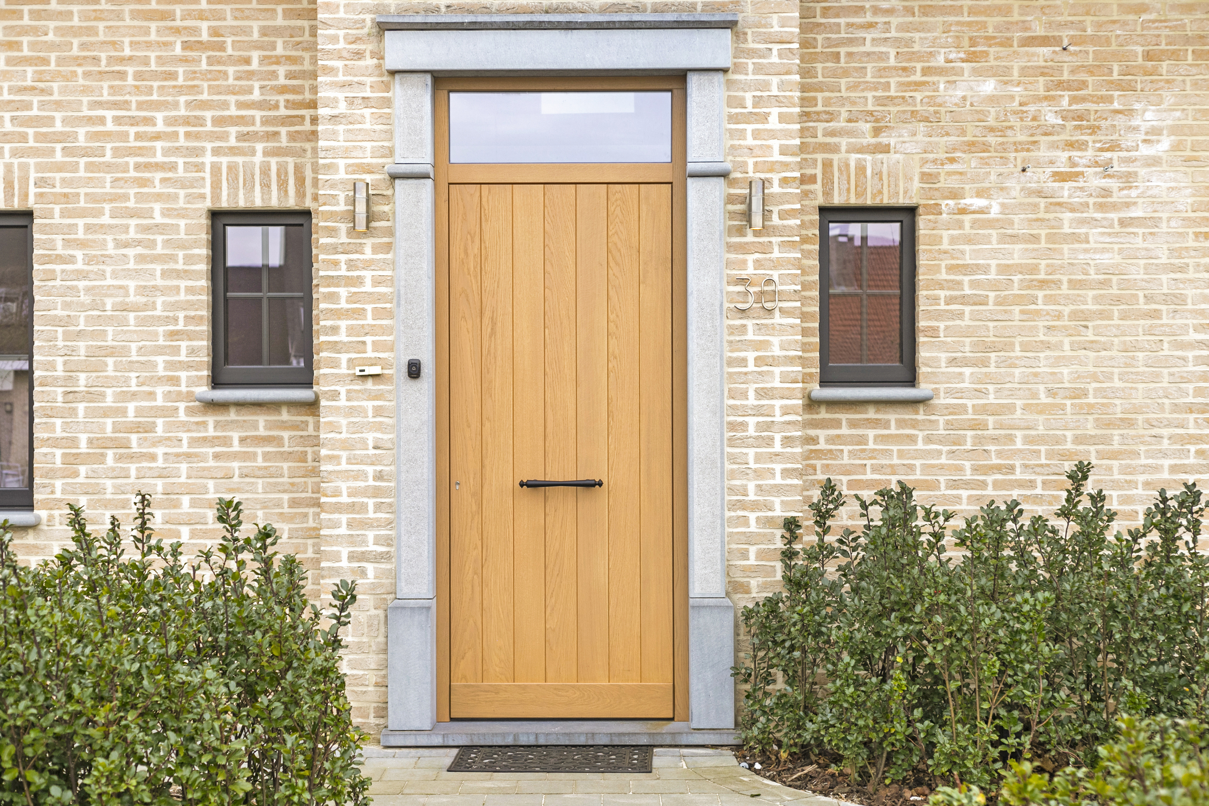Façade en briques rénovée avec une porte d’entrée en chêne au style chaleureux et champêtre, des châssis noirs mats, une imposte et de petites fenêtres fixes et oscillo-battantes.