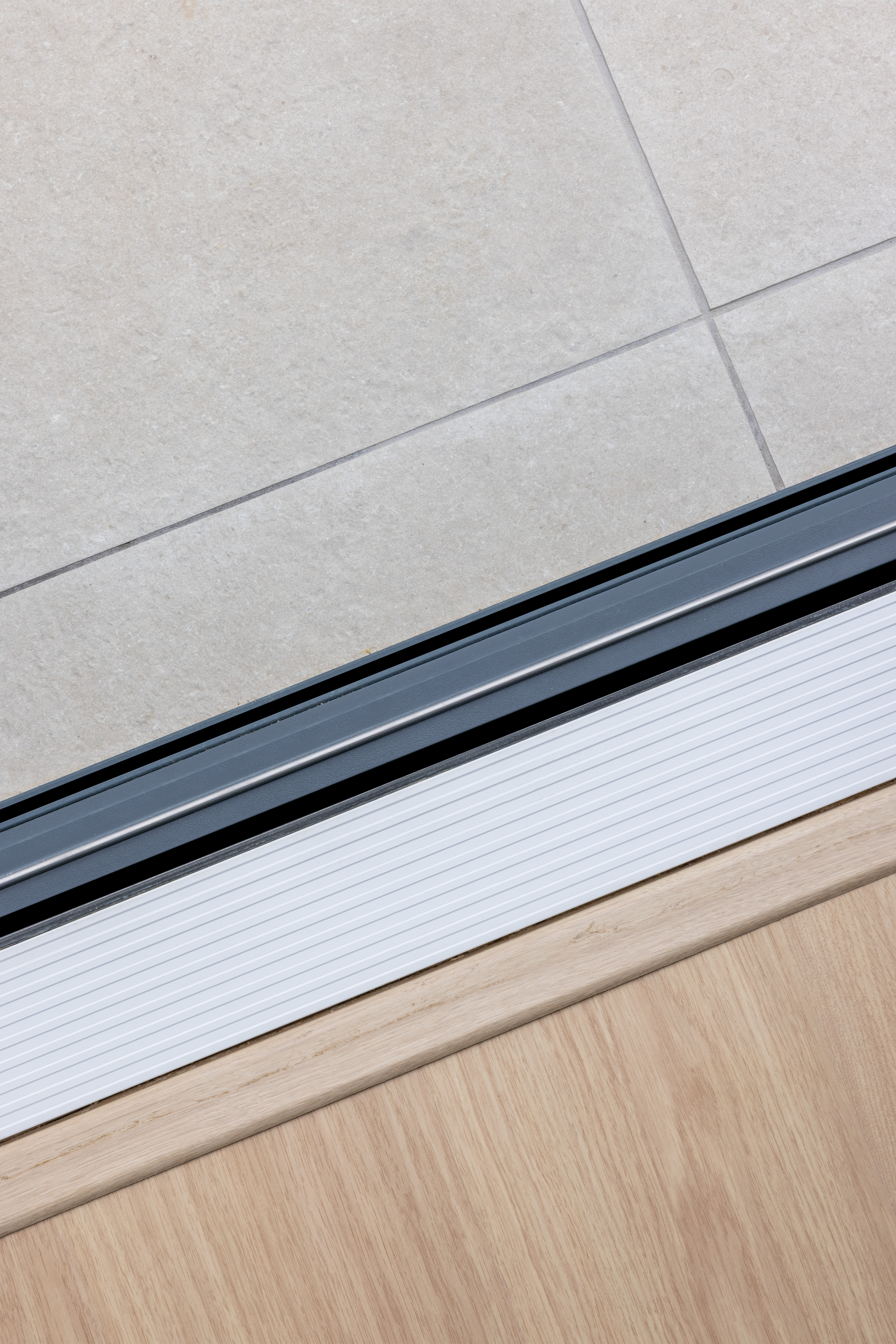 Detail van een modern ALU Visoglide Monorail-schuifraam in mat verkeerswit, met zichtbare rail tussen lichte vloertegels en een houten vloer tijdens een renovatie.