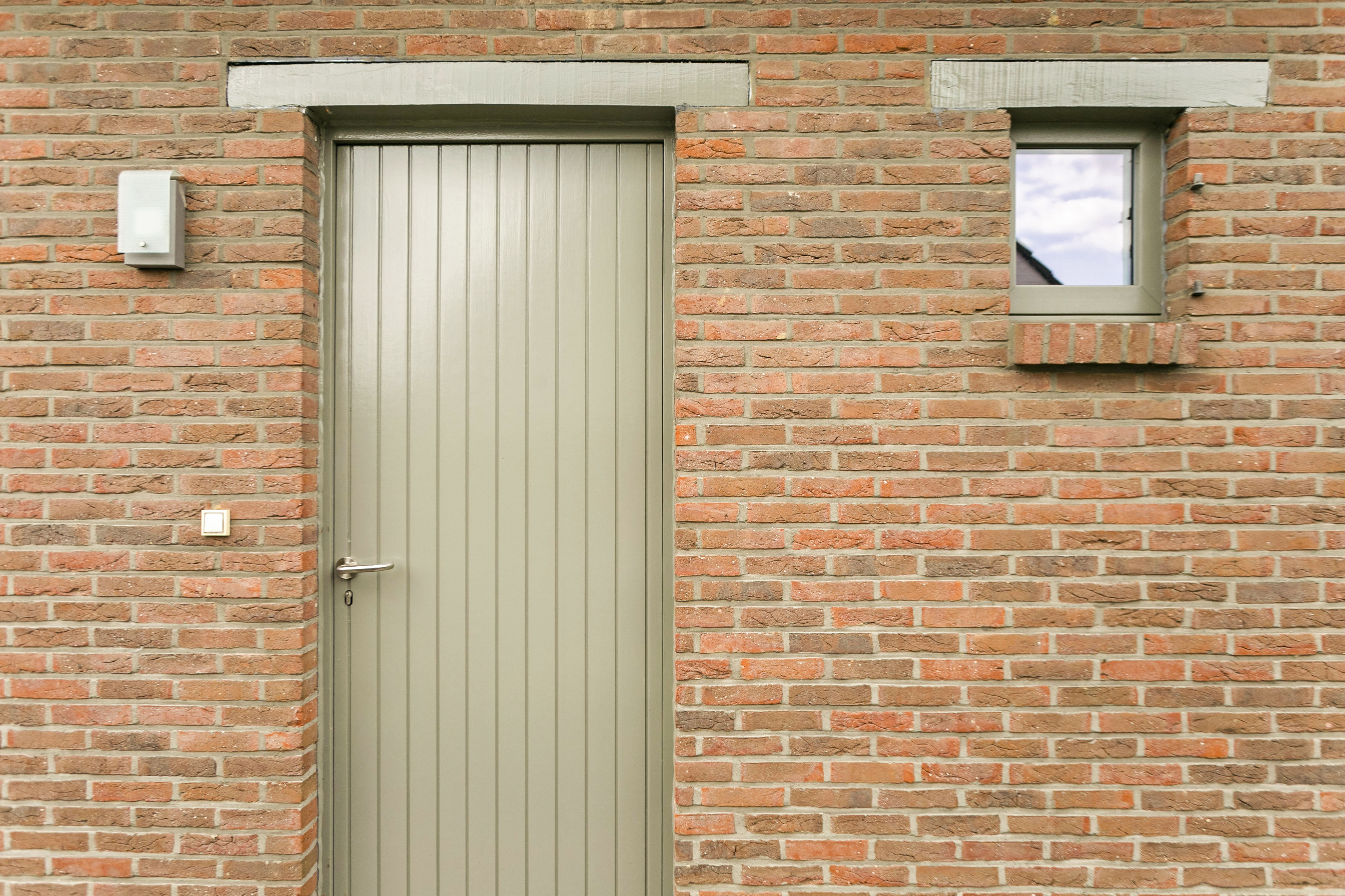 Betongrijs vast raam met verticale profilering in een gevel van rode baksteen, naast een klein raam.