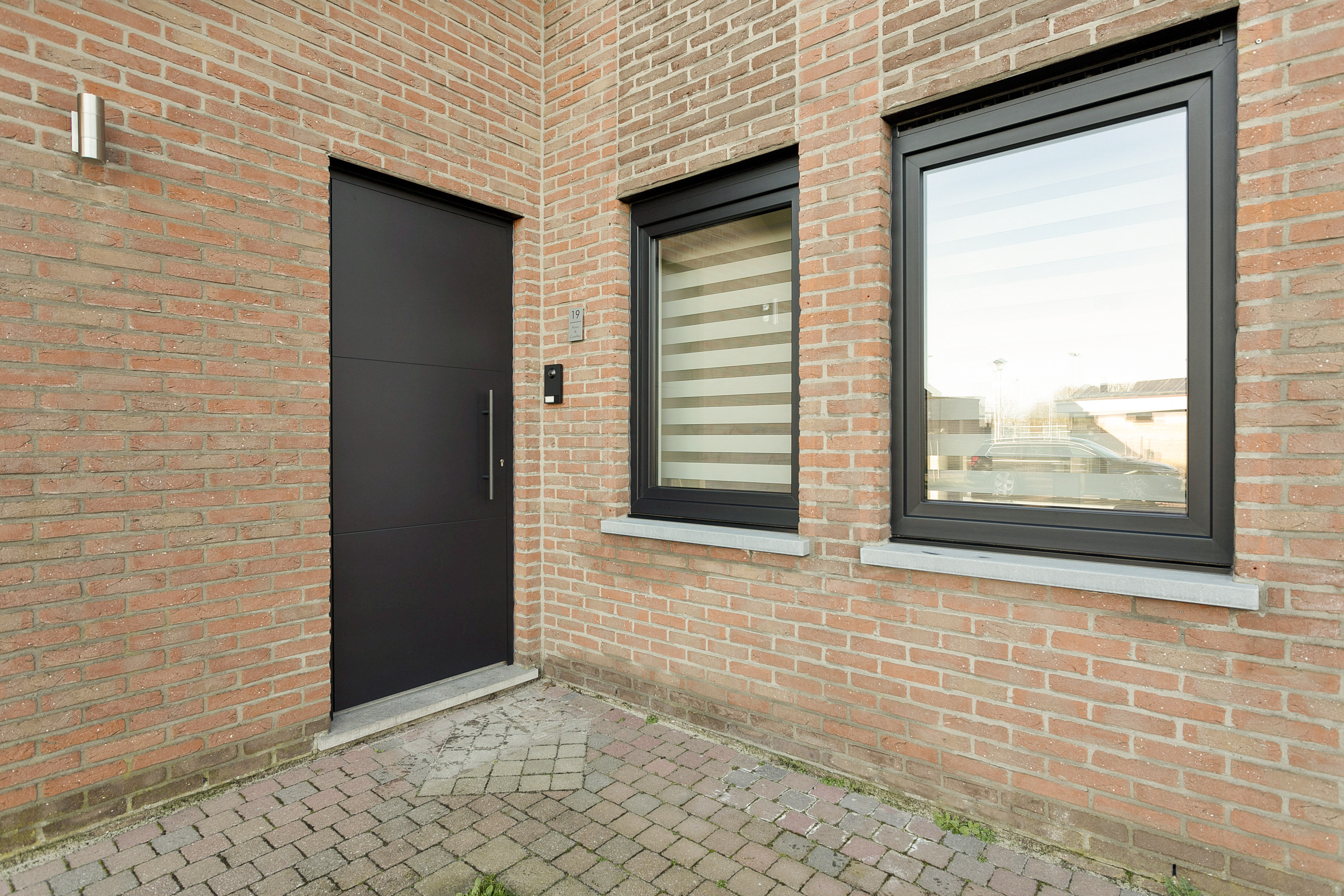 Zwart mat PVC-draai-kipraam in een gerenoveerde bakstenen gevel, naast een zwarte deur en een betegeld toegangspad.