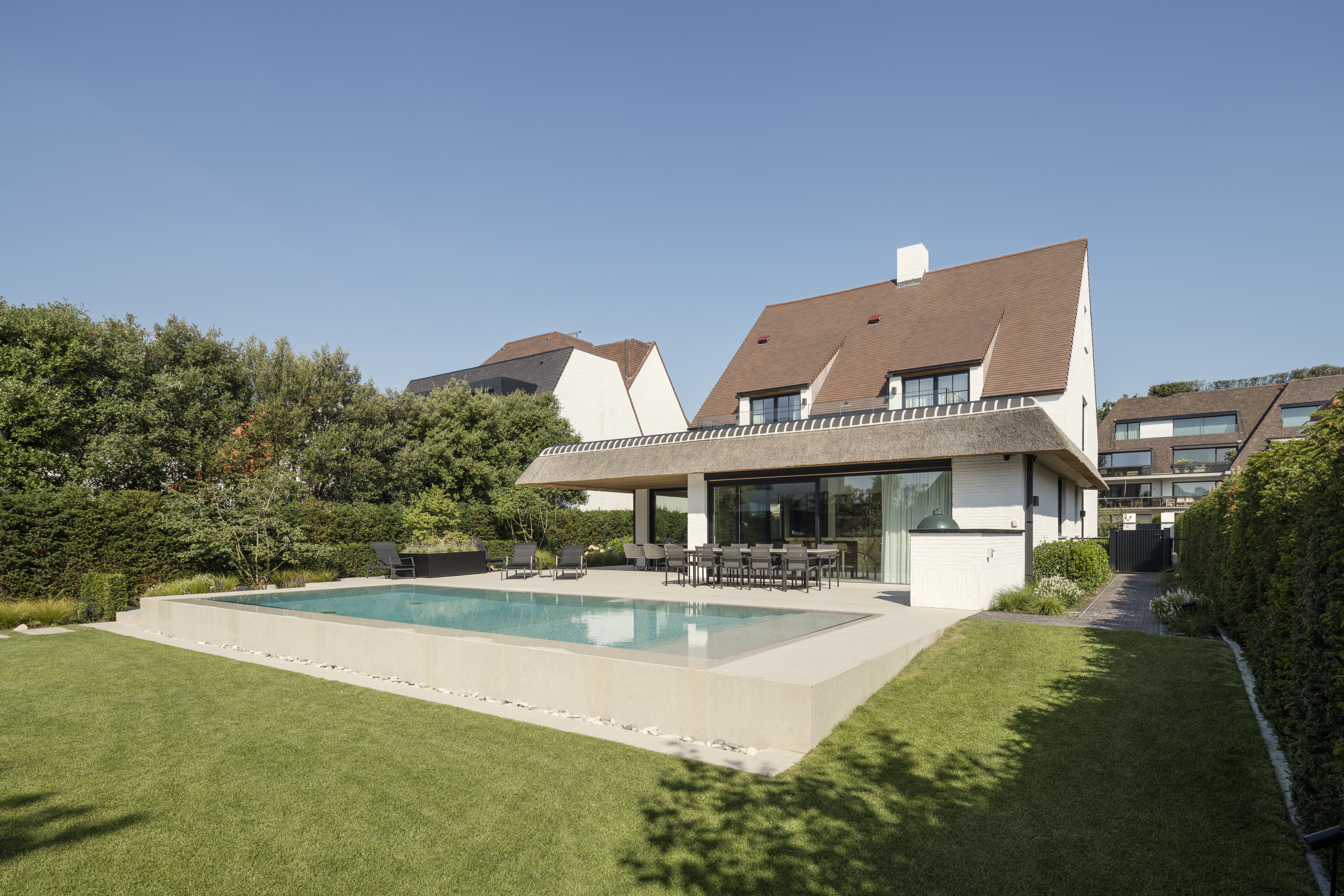 Moderne woning met groot terras, kamerhoge raampartijen en een rechthoekig zwembad in een verzorgde tuin onder een blauwe hemel.