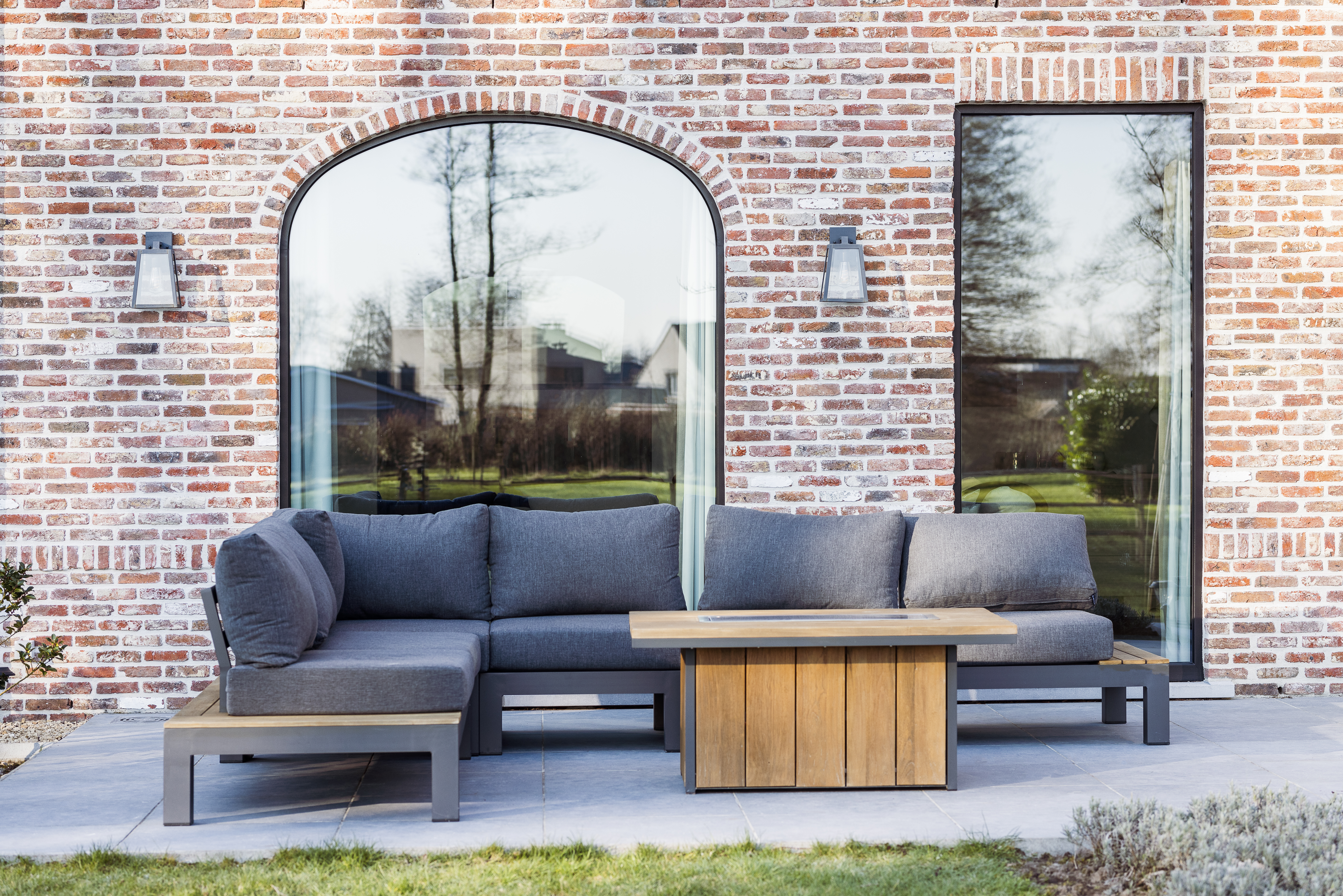 Bakstenen gevel met een vast, rondboogvormig raam en een smal rechthoekig raam met matzwarte Impakt PLUS-profielen; ervoor staan een loungeset en tafel.