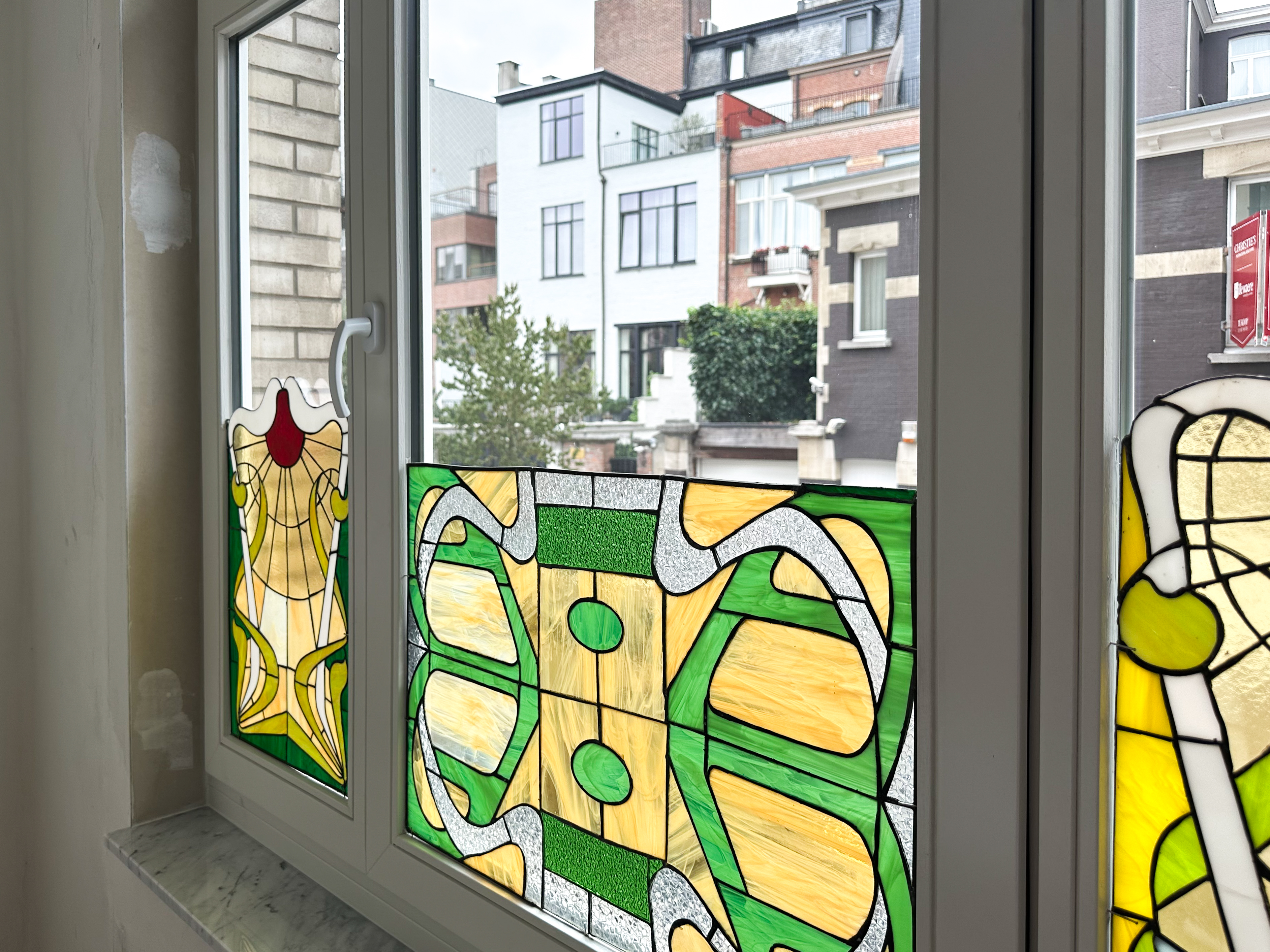Gerestaureerd vast raam en draai-kipraam in verfijnde klassieke stijl, met grijs afgelakte raamprofielen en kleurrijk glas in een gesloten bebouwing in Brabant.