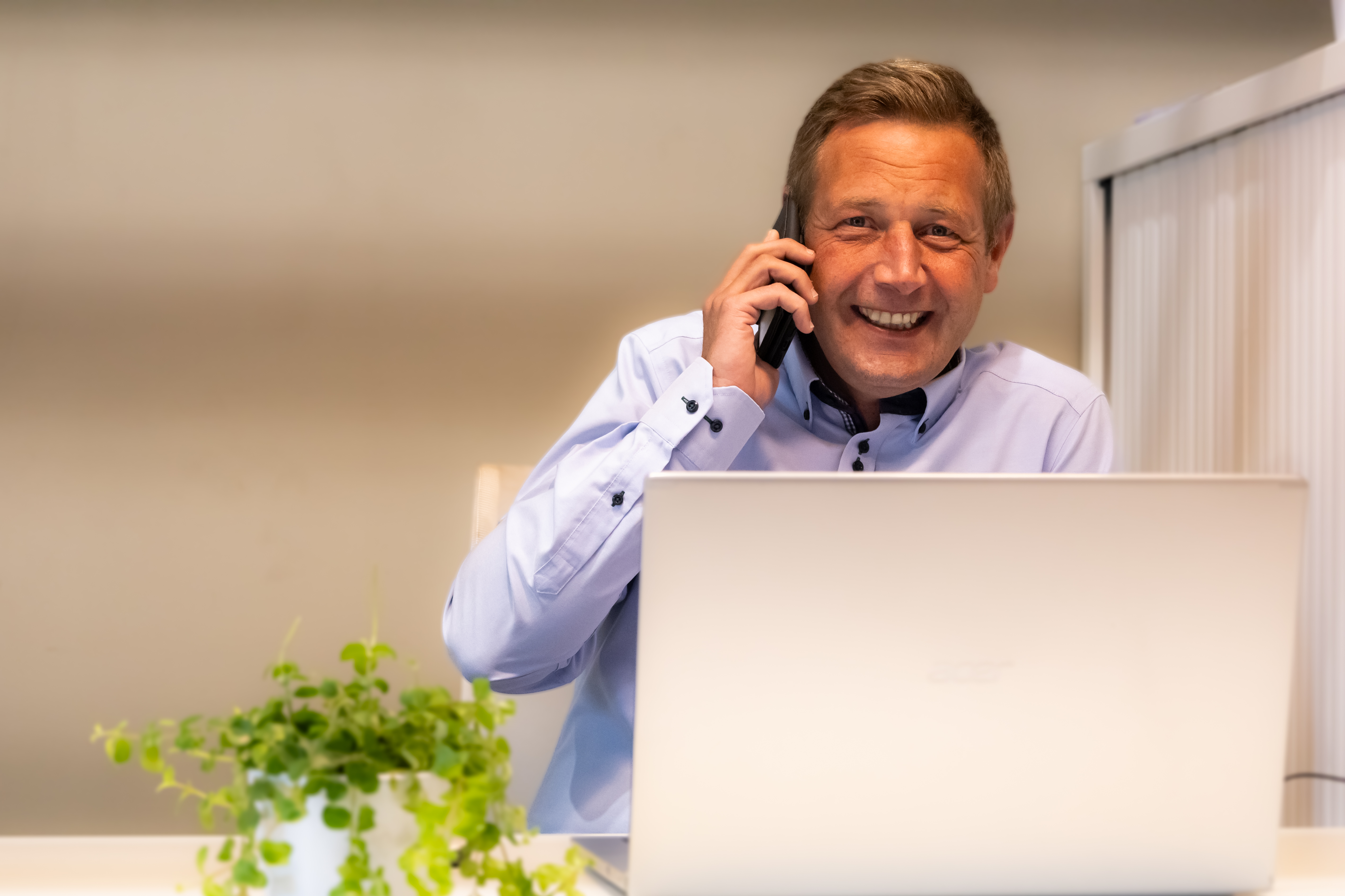 Een glimlachende persoon belt met een smartphone terwijl hij achter een laptop zit, in een lichte binnenruimte met een groene plant.
