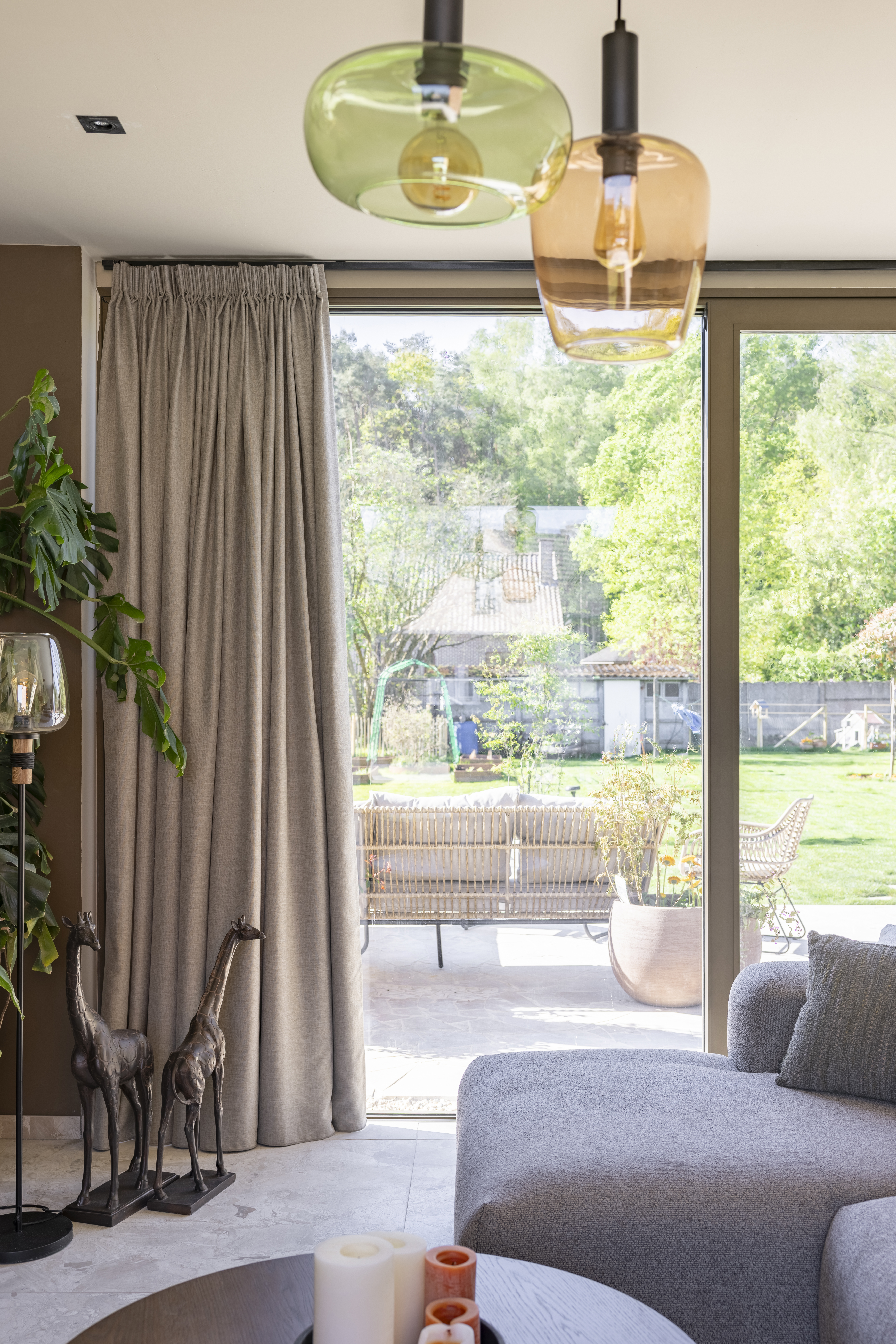 Interieur met een groot ALU Visoglide Monorail-schuifraam in C33-bronsanodisatie, met uitzicht op een groene tuin, naast gordijnen en een zithoek.