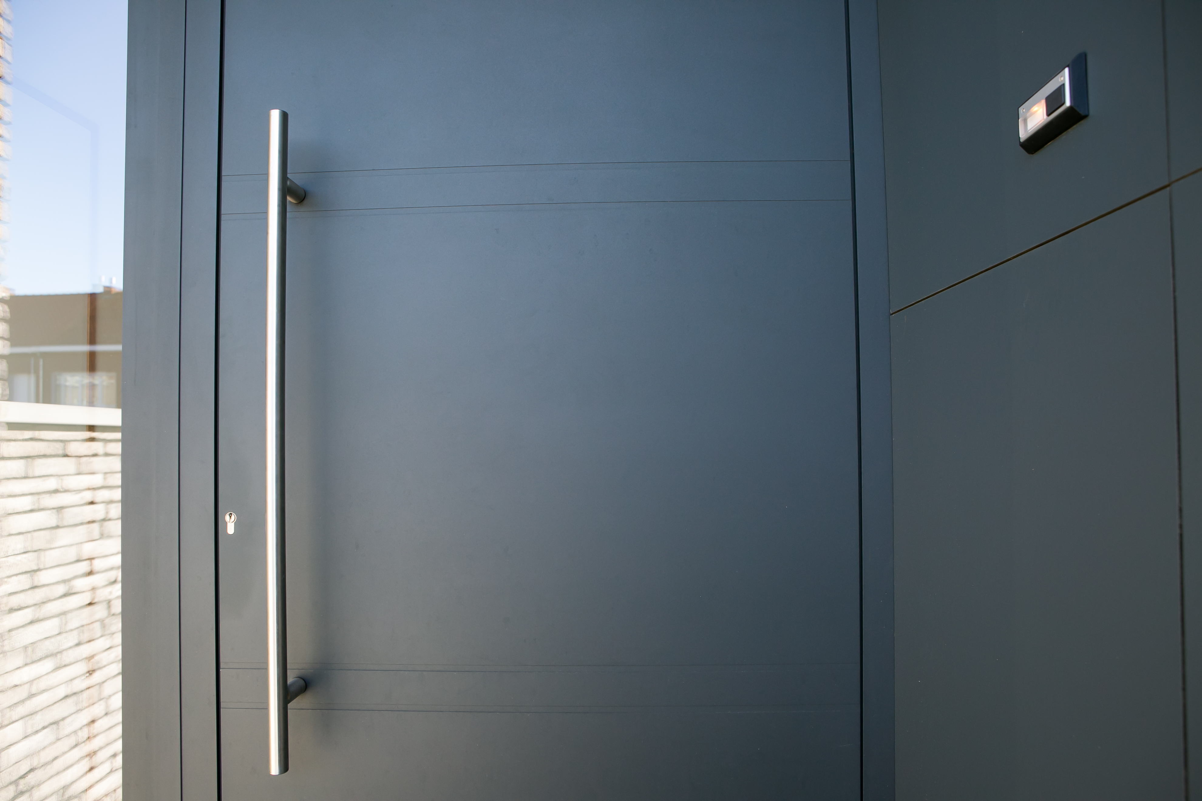 Moderne donkergrijze aluminium voordeur met lange verticale handgreep en zijlicht, geplaatst in een strakke gevel van een nieuwbouwwoning.