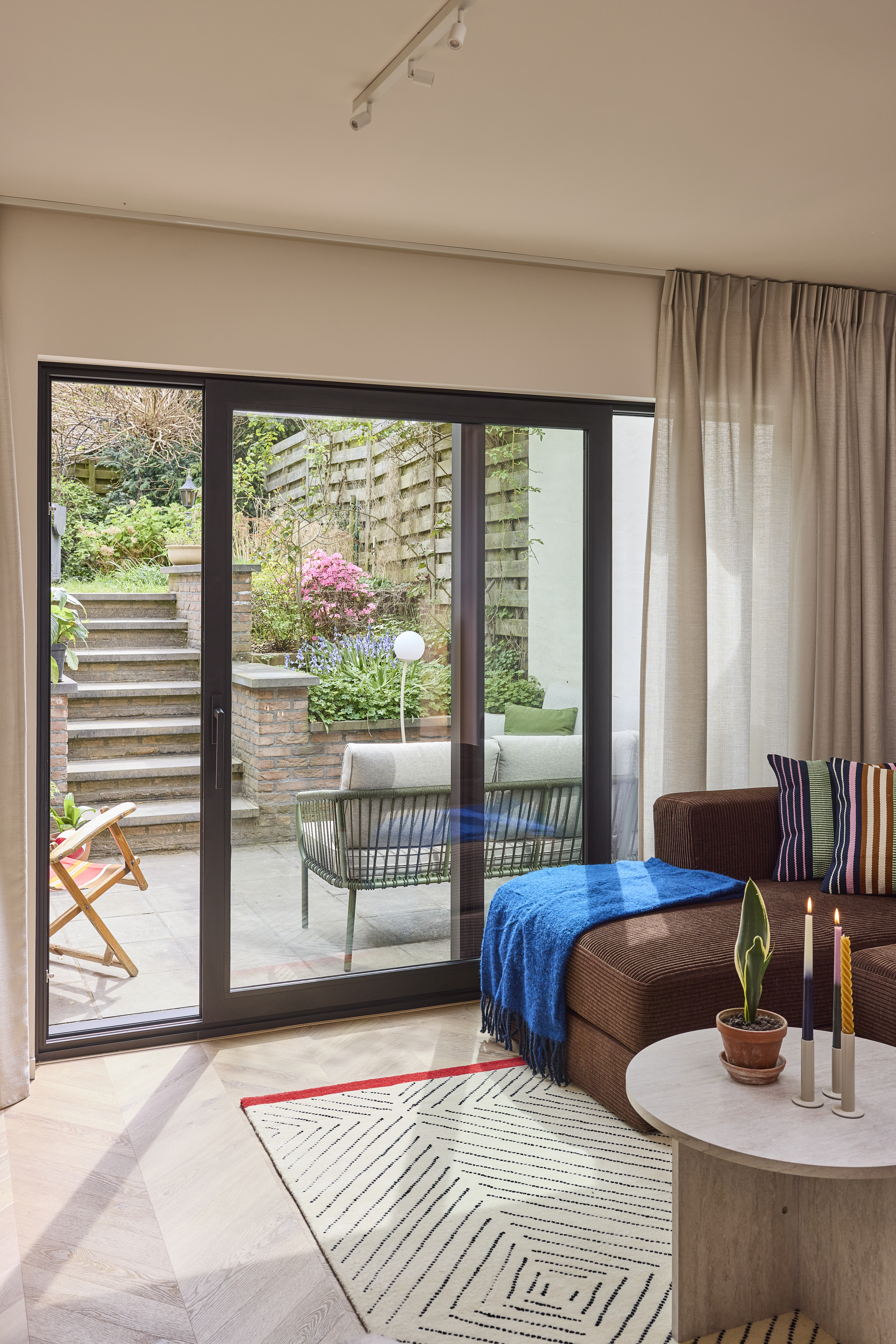 Lichte woonkamer met een signaalzwart ALU Visoglide Monorail-schuifraam naar een groene terraszone met trap, naast een sofa, salontafel en vloerkleed.