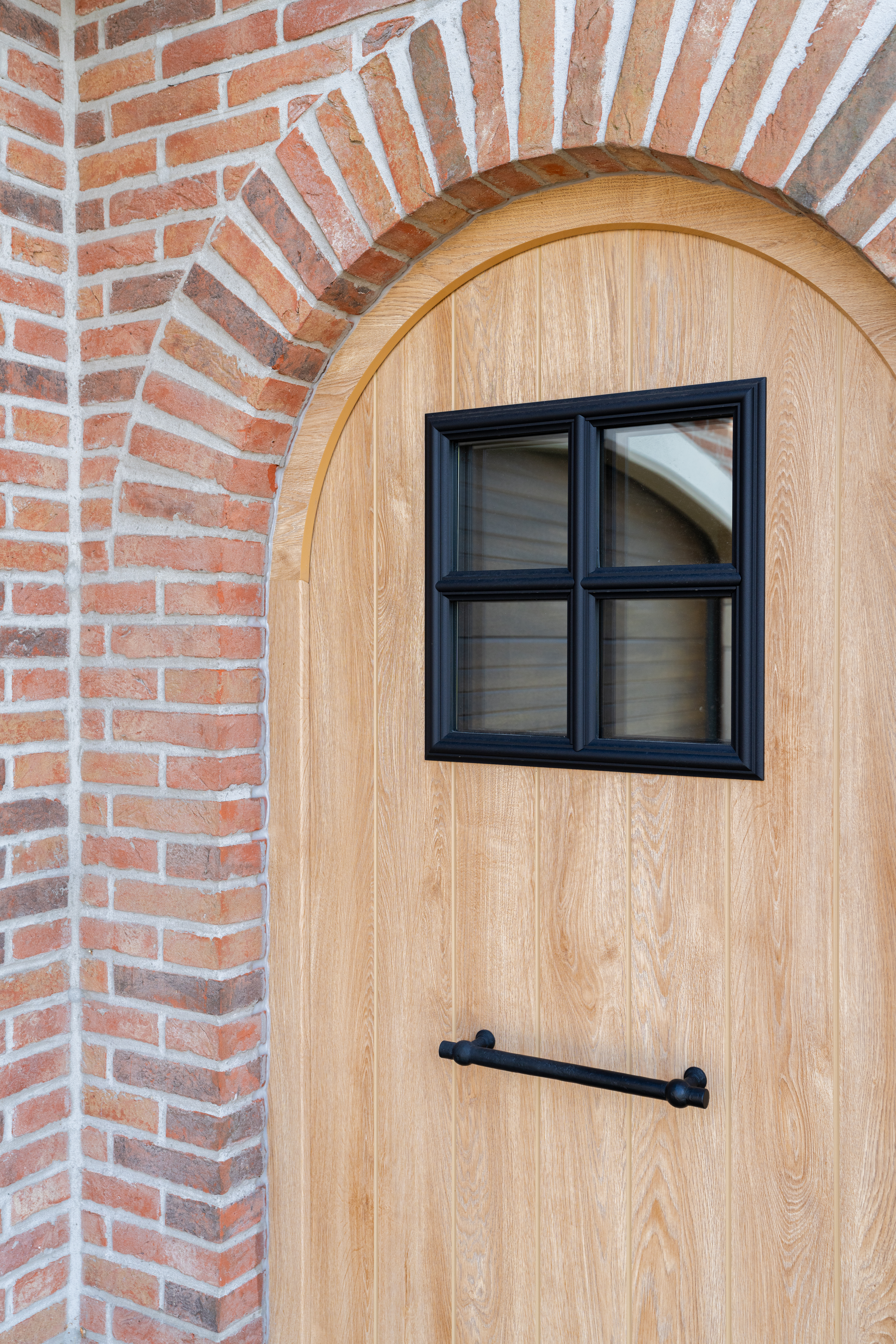 PVC-deurpaneel in natuurlijke eiklook met een klein zwart raam en horizontale handgreep, omgeven door een boog van rode bakstenen.